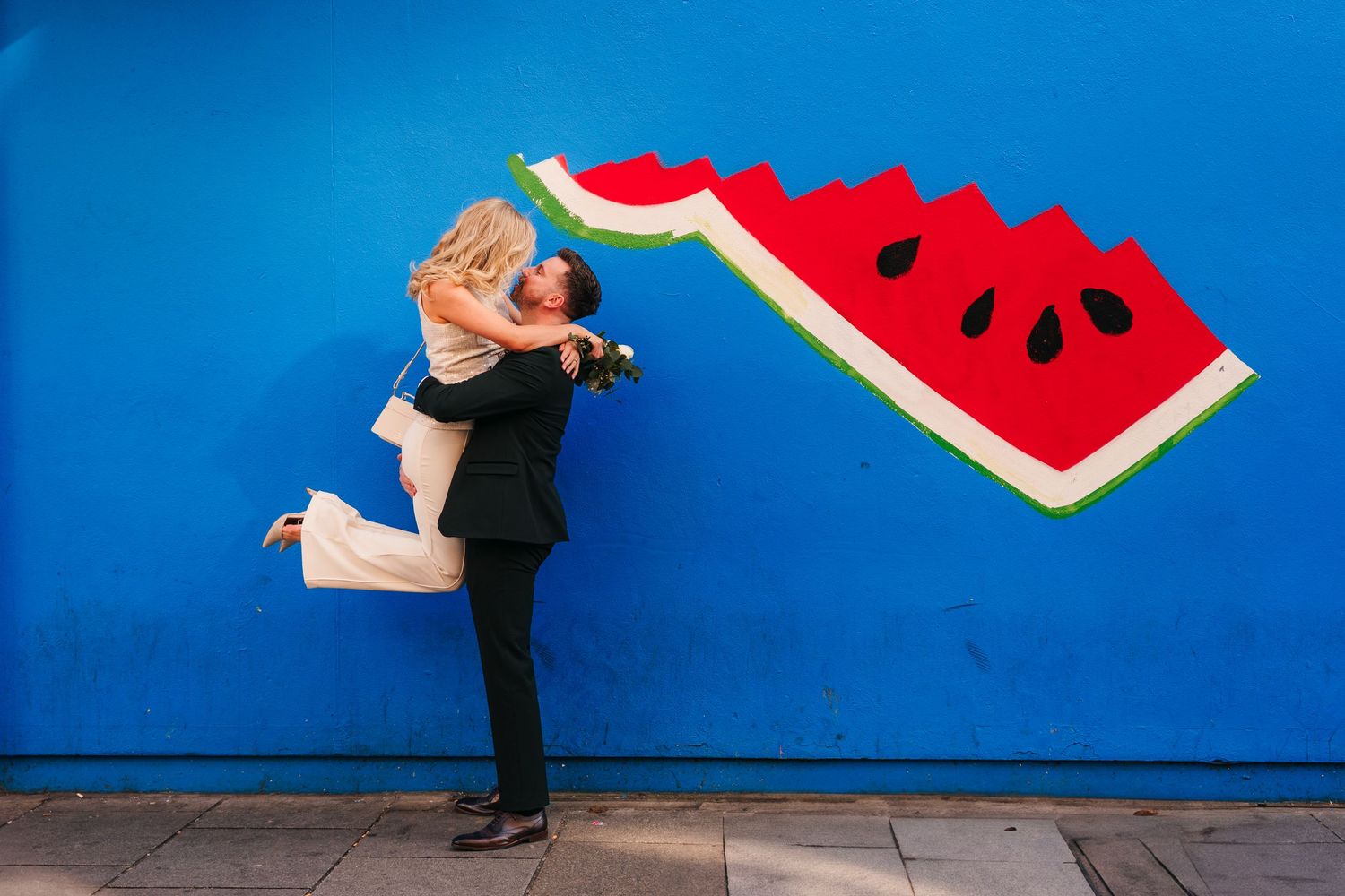 A couple embraces in front of a bright blue wall with a painted watermelon slice mural. City Hall Dublin wedding photogrAPher