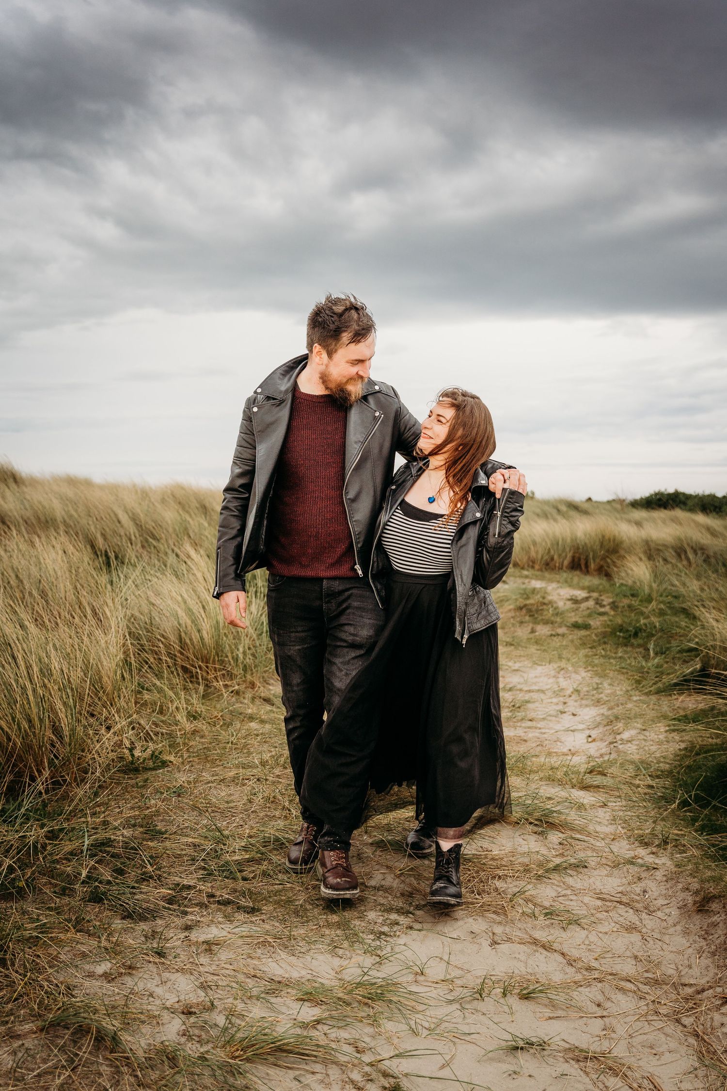 Couple walking together on sandy coastal path through beach grass under moody sky. engagement PHOTOGRAPHER Dublin