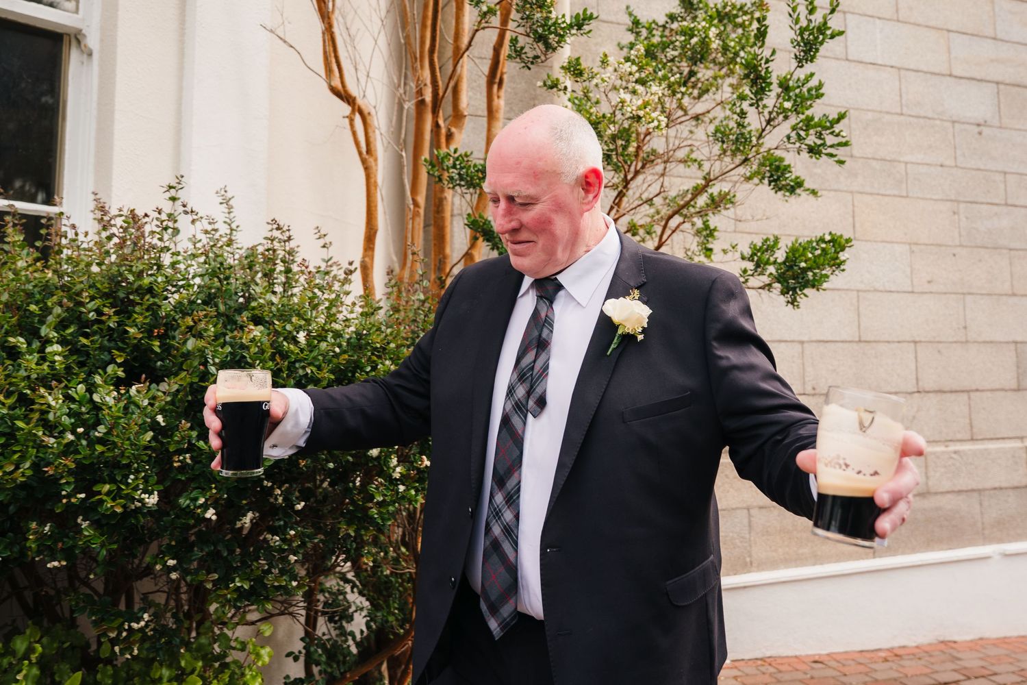 An elegantly dressed man in a black suit gestures enthusiastically outside a building. Portmarnock Hotel Wedding PHOTOGRAPHER Dublin
