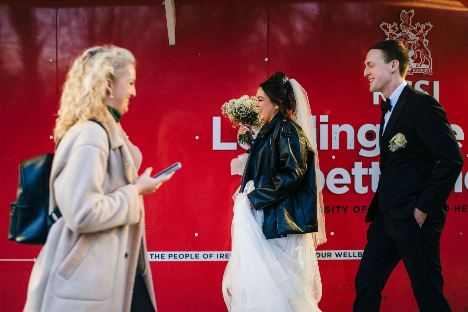 A couple in wedding attire interact with someone near a red wall outdoors. Wedding PHOTOGRAPHER Dublin