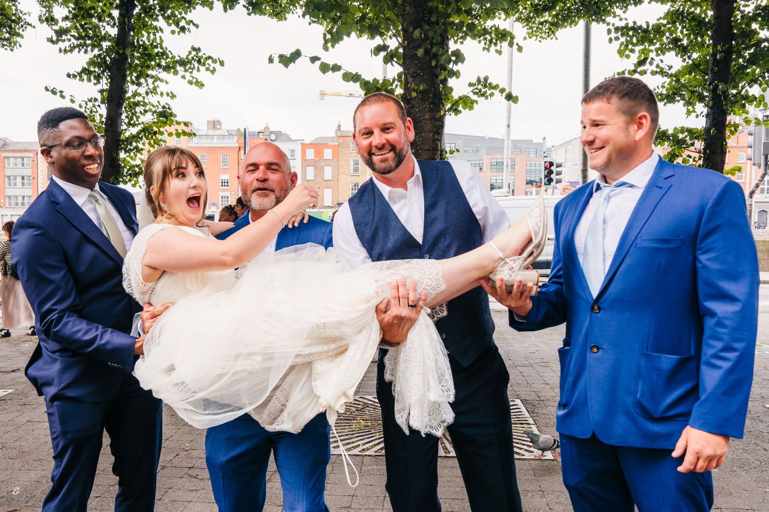 Wedding party lifts the bride in a playful group photo outdoors under tree canopy. PHOTOGRAPHER Dublin Ireland Wedding