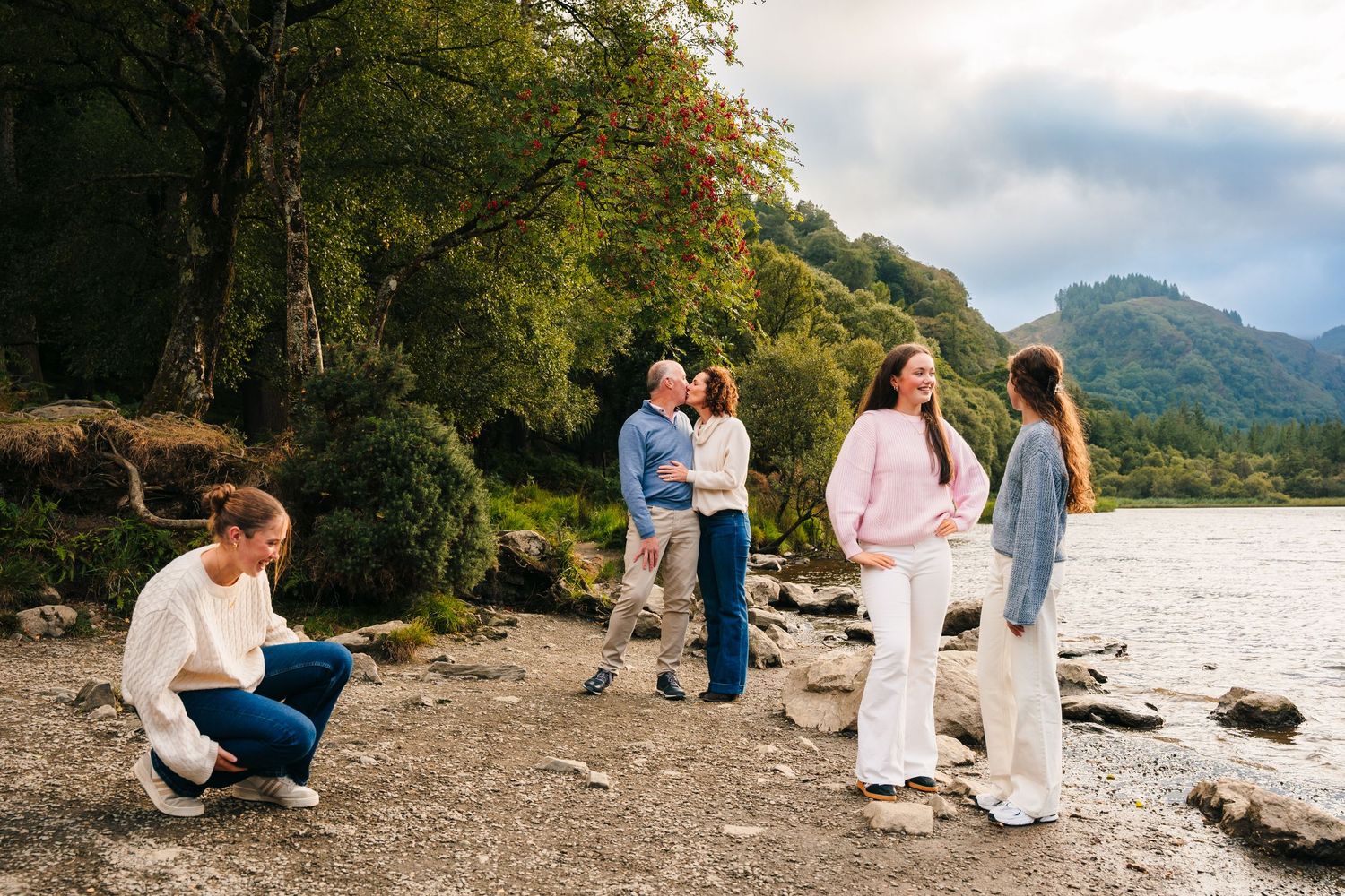 Family photographer IrelanA lakeside photoshoot scene with people in casual attire gathered on a rocky shore with mountains in the distance.