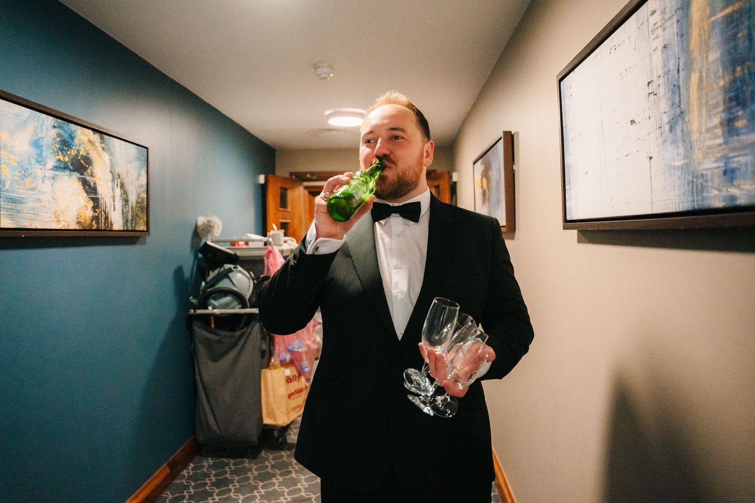 A person in a black tuxedo drinking champagne while holding wine glasses in an elegant hallway. Wedding Photography Trim Castle Ireland