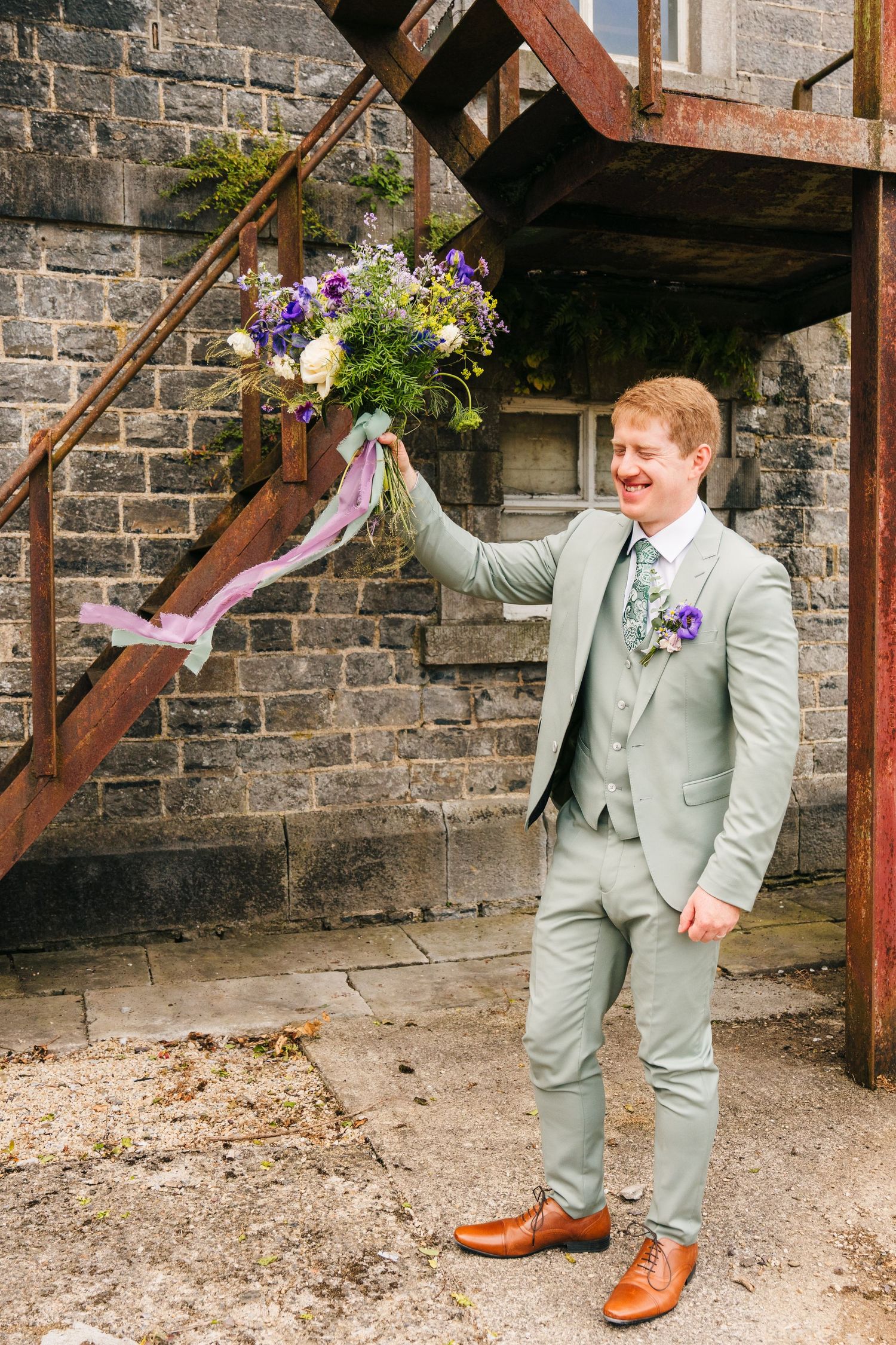 A groom in a light gray suit holds up a purple floral bouquet against a rustic brick wall with wooden stairs.Millhouse slane wedding photogr