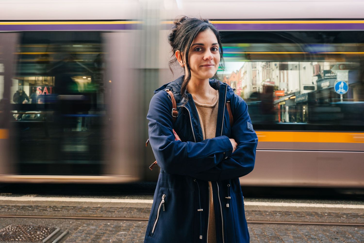 Person in a blue denim jacket stands in front of a blurred urban background with passing traffic and city lights. professional headshots dub