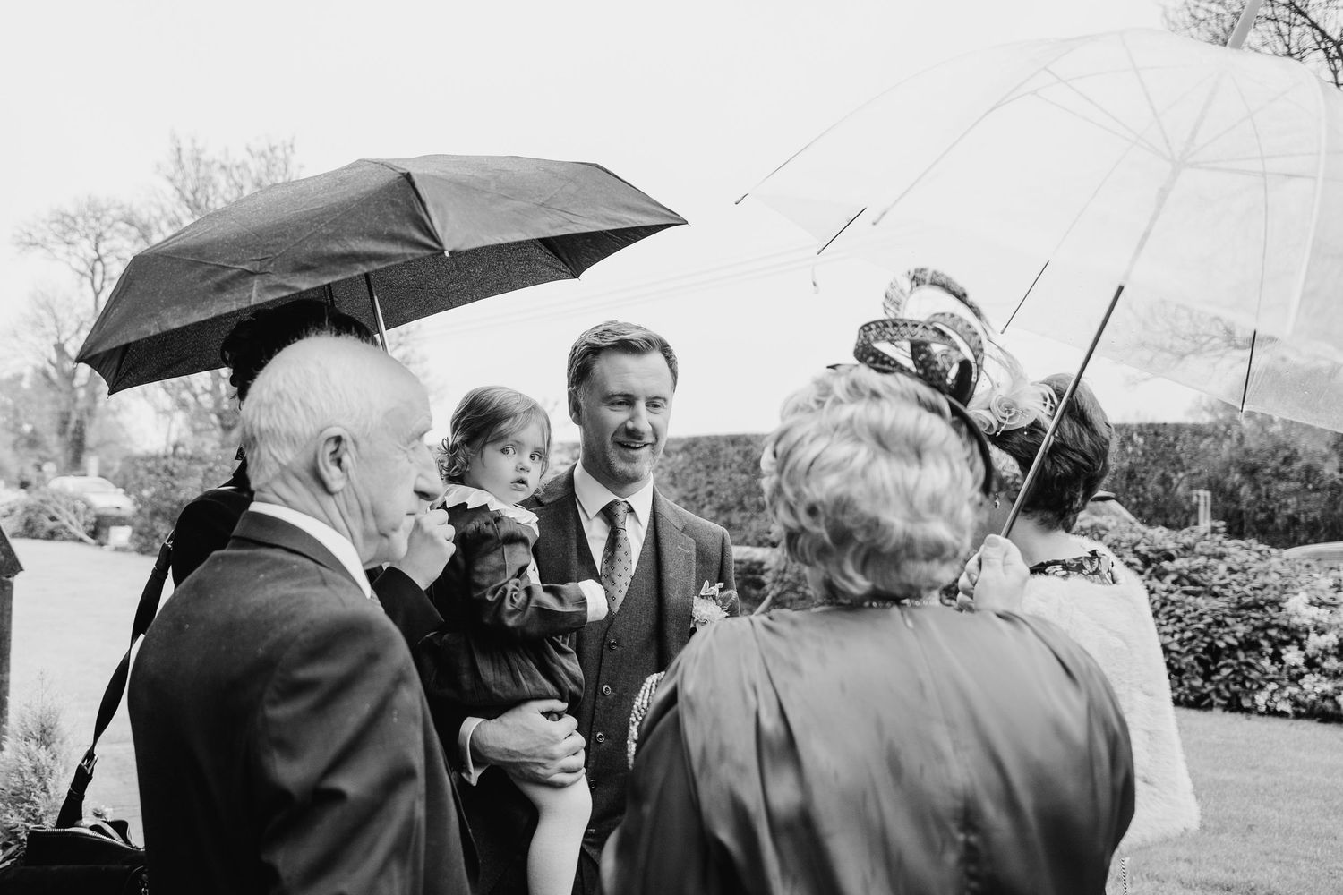 Wedding guests huddle under black umbrellas during an outdoor rainy celebration.