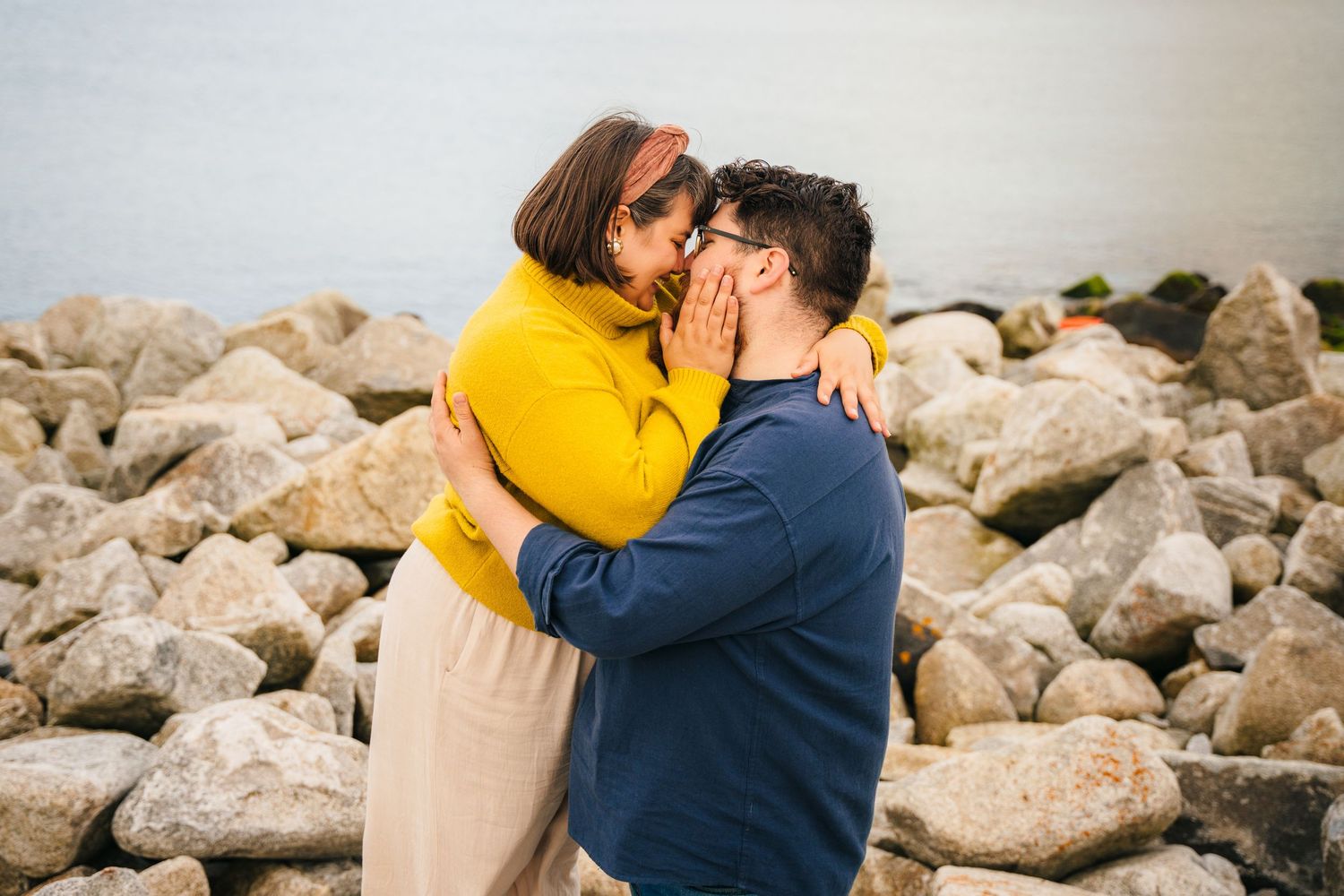 A couple shares a tender kiss while standing on a rocky beach shore, one person wearing a bright yellow sweater. PHOTOGRAPHER Dublin ireland