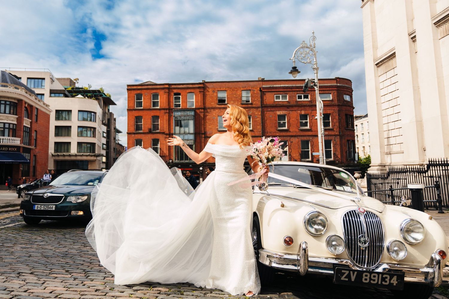 Elegant wedding day scene with flowing white dress and buildings against a blue sky in an urban setting.