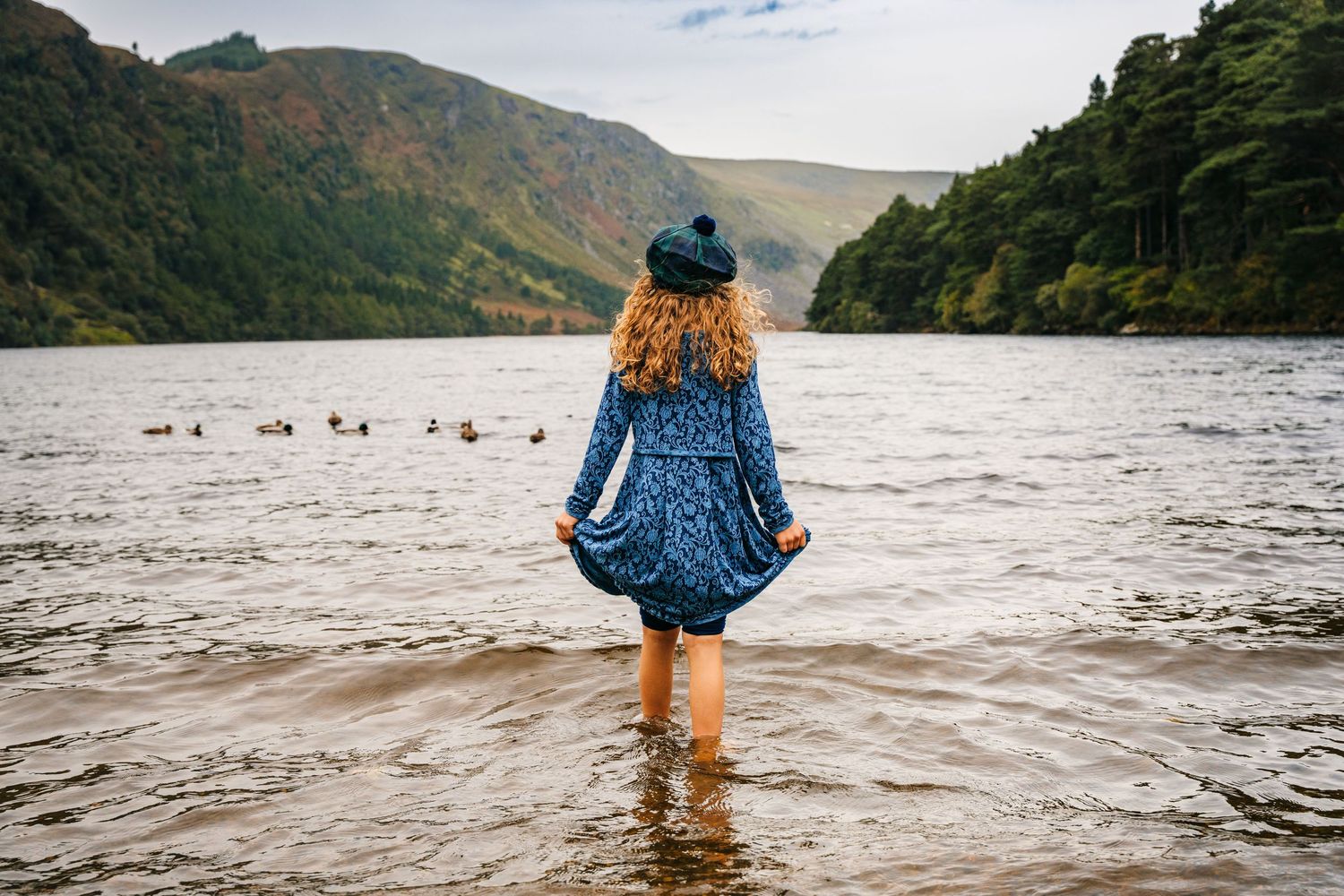 A person wearing a blue dress stands in shallow lake water with mountains and forest in the background.Family photogrAPHER DUBLIN