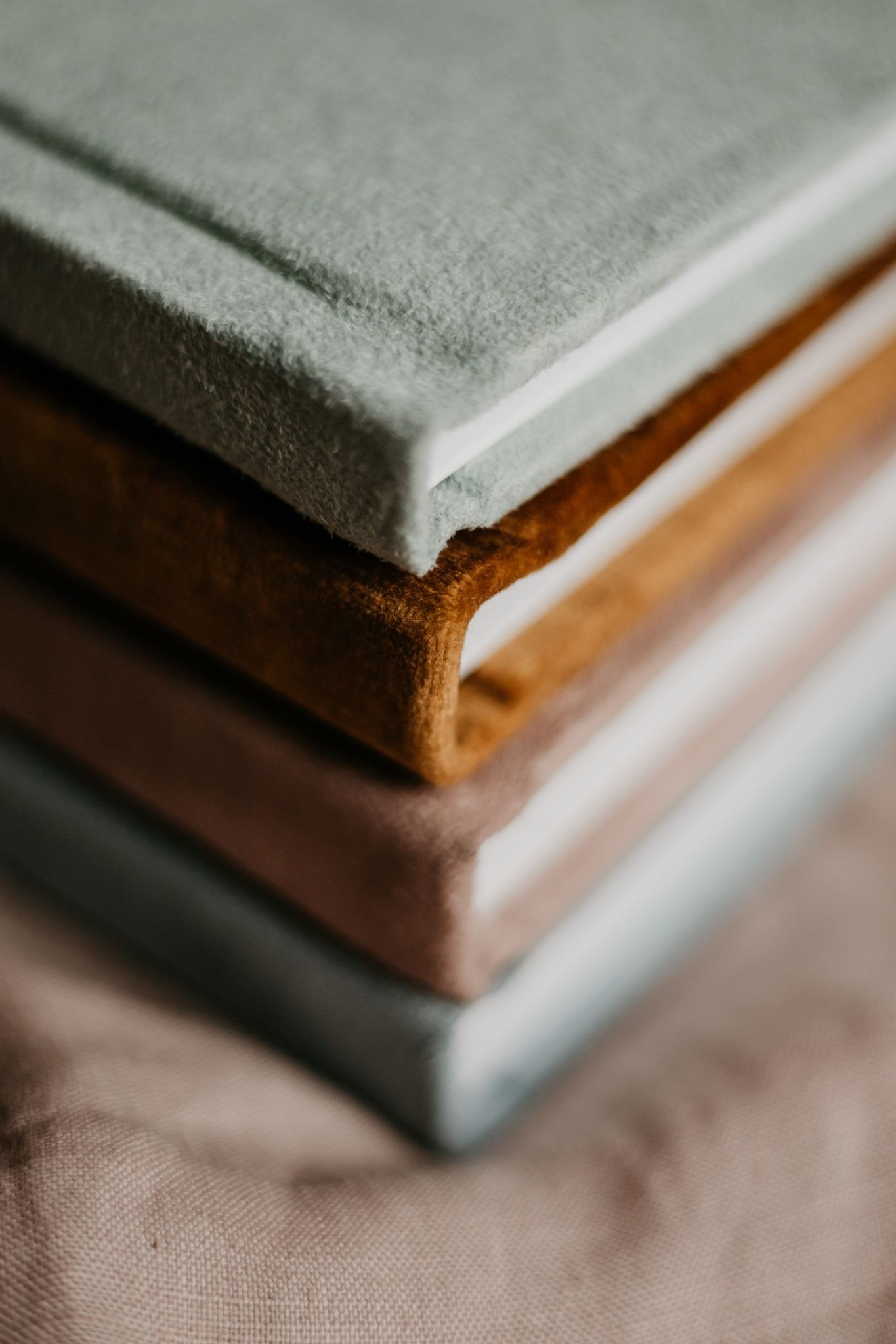 Stack of four fabric-covered books in sage green, tan, blush pink, and gray tones on a linen surface.