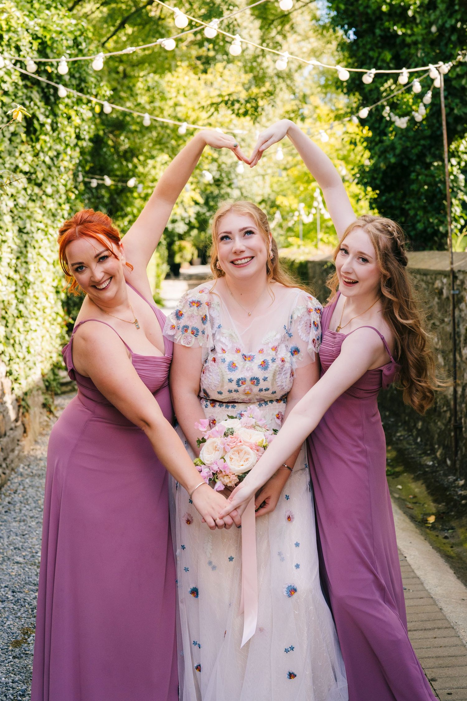 Three wedding party members pose together wearing a floral embellished gown and matching mauve bridesmaid dresses. . Village Barn Tyrrellspa