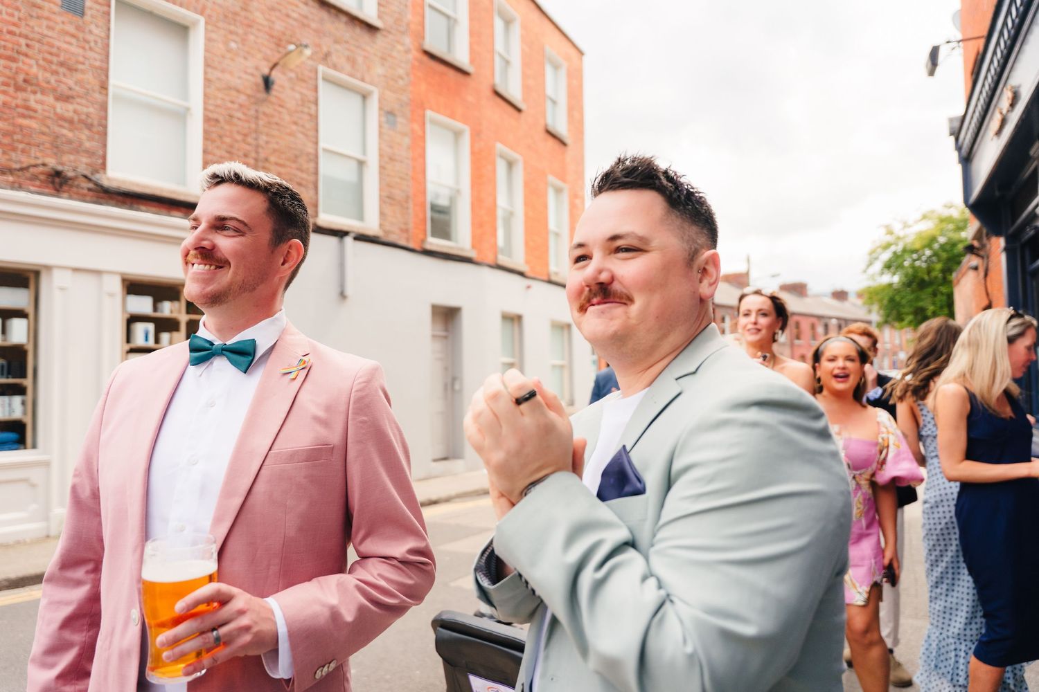 People in formal attire gather at a street celebration wearing pink and gray suits. Same sex wedding PHOTOGRAPHER Dublin