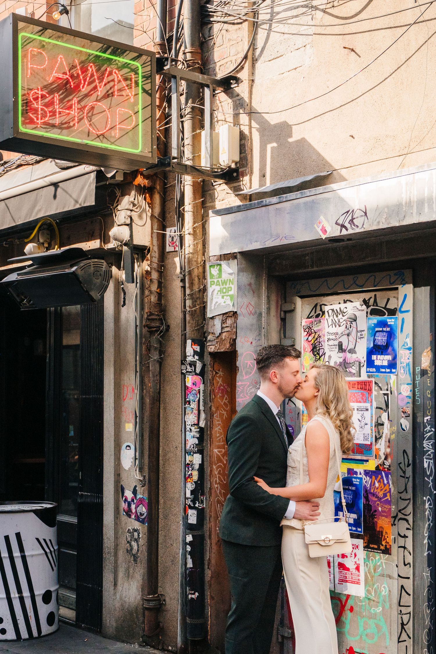 A romantic moment shared between a couple in front of a neon-lit urban wall covered in street art and posters. City Hall Dublin wedding phot