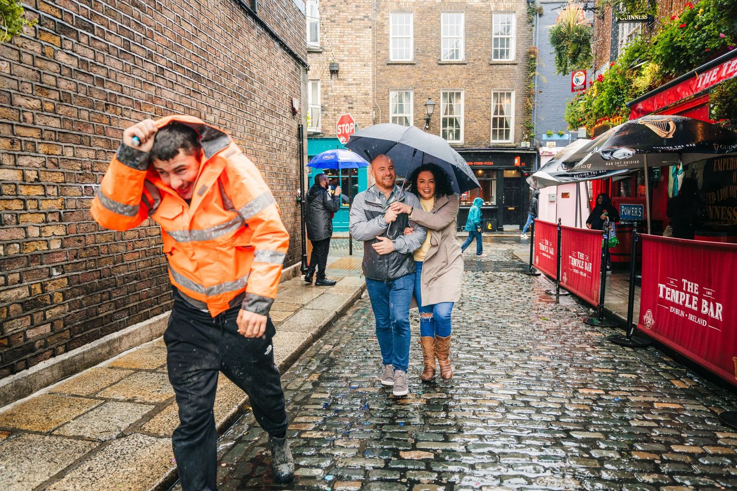 A couple poses romantically on a rainy cobblestone street in Edinburgh with red umbrellas and brick buildings. Proposal Dublin Photographer
