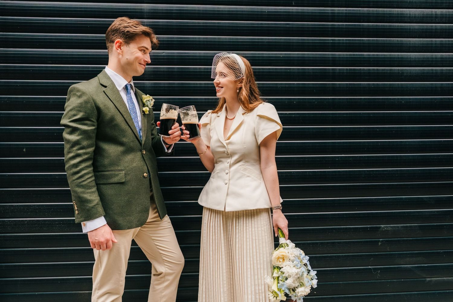 A couple in wedding attire toasting with red wine, the groom in a green blazer and bride holding a white bouquet.