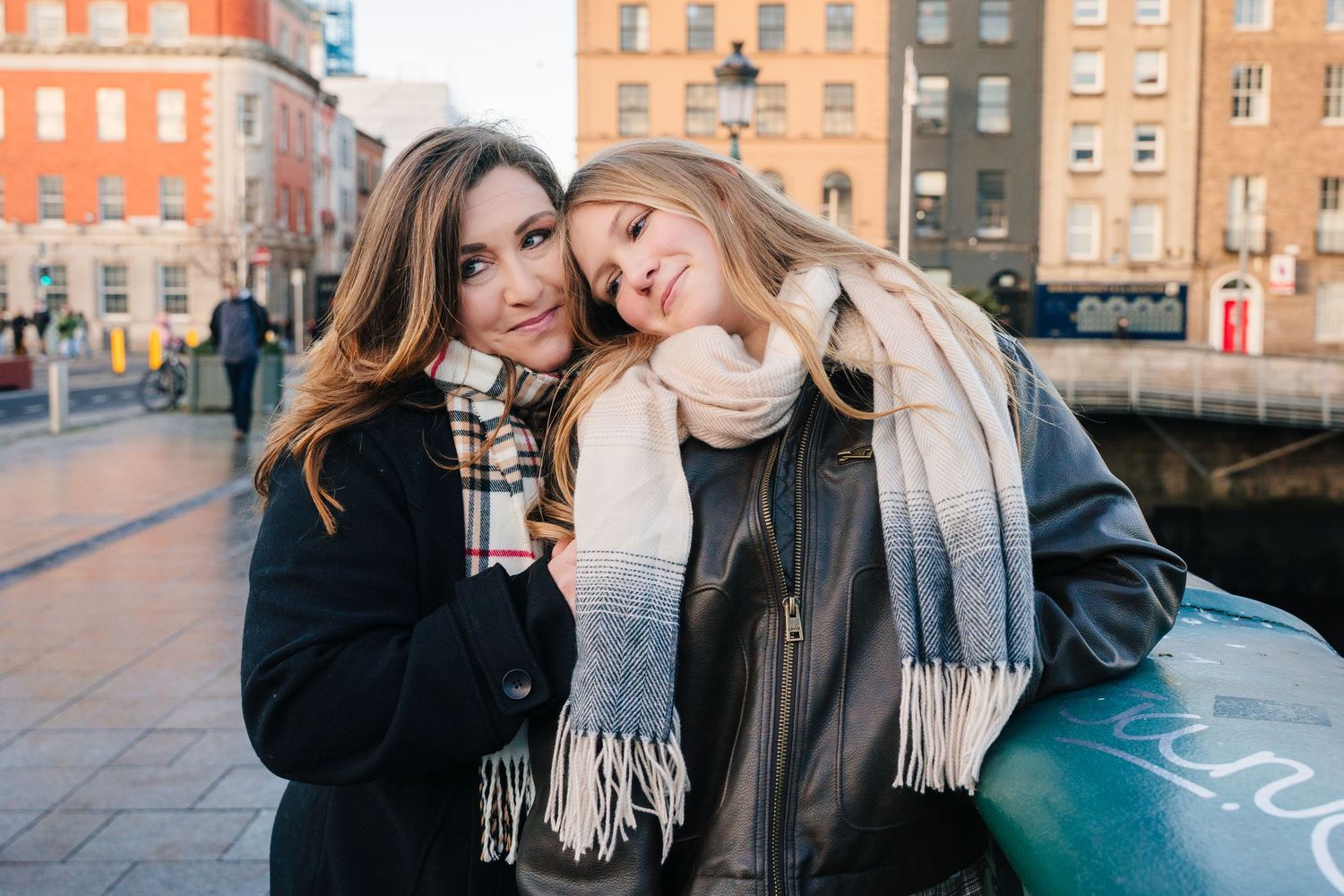 Two friends in winter coats and scarves lean together while enjoying a sunset stroll along an urban canal.