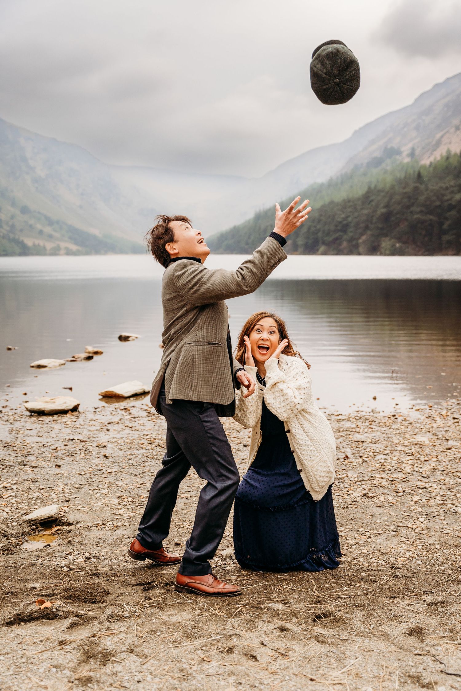A couple sits playfully by a serene mountain lake, tossing a hat in the air against a misty landscape backdrop. photogrAPHER DUBLIN IRELAND