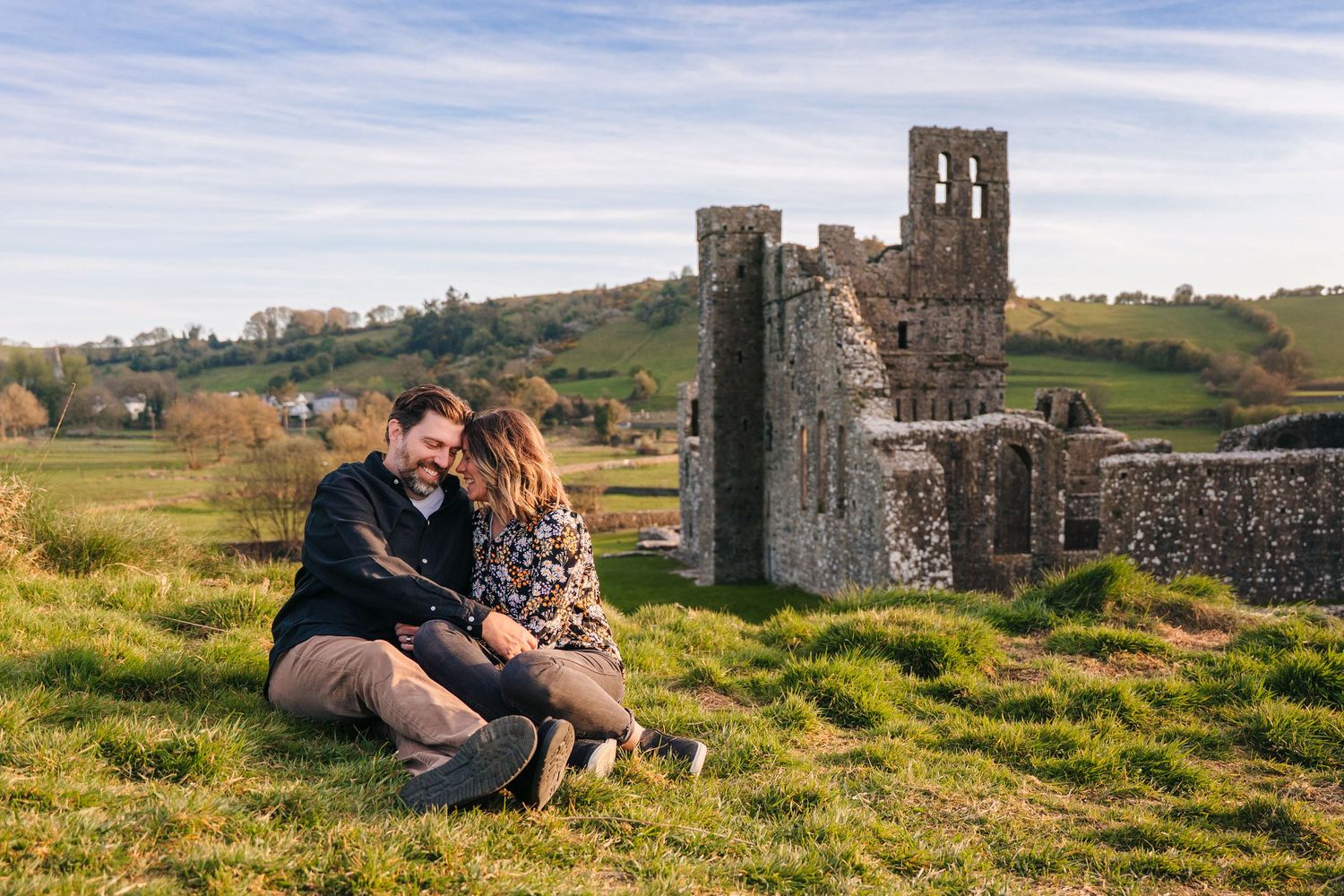 A couple sits together on a grassy hill near ancient stone ruins during golden hour. PHOTOGRAPHER Dublin ireland