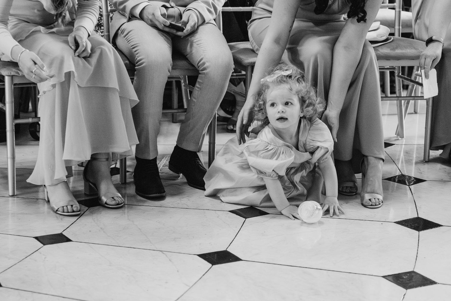 Black and white photo of a small child sitting on checkered floor during wedding reception.  city hall dublin wedding photos