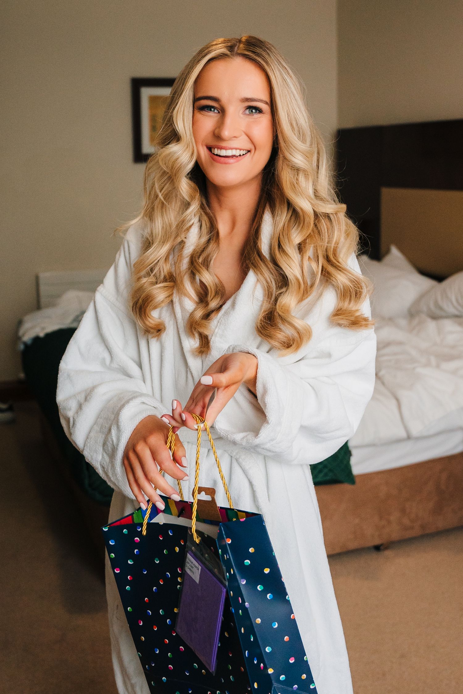 Woman in white bathrobe holds shopping bags while smiling in a hotel room. Wedding Photography Trim Castle Ireland