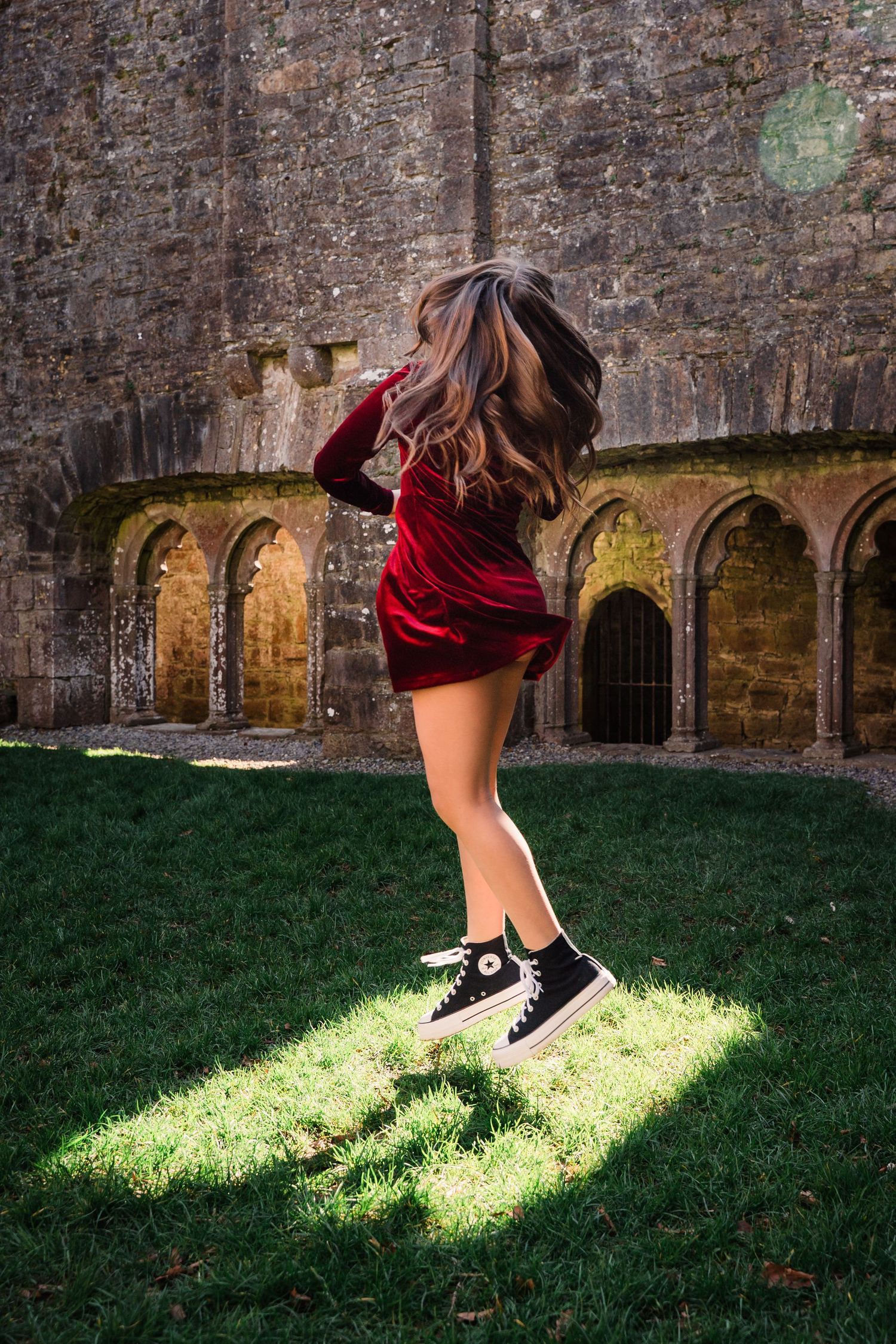 Person in red velvet dress and sneakers jumps joyfully in front of ancient stone castle archways. Family PHOTOGRAPHER Dublin