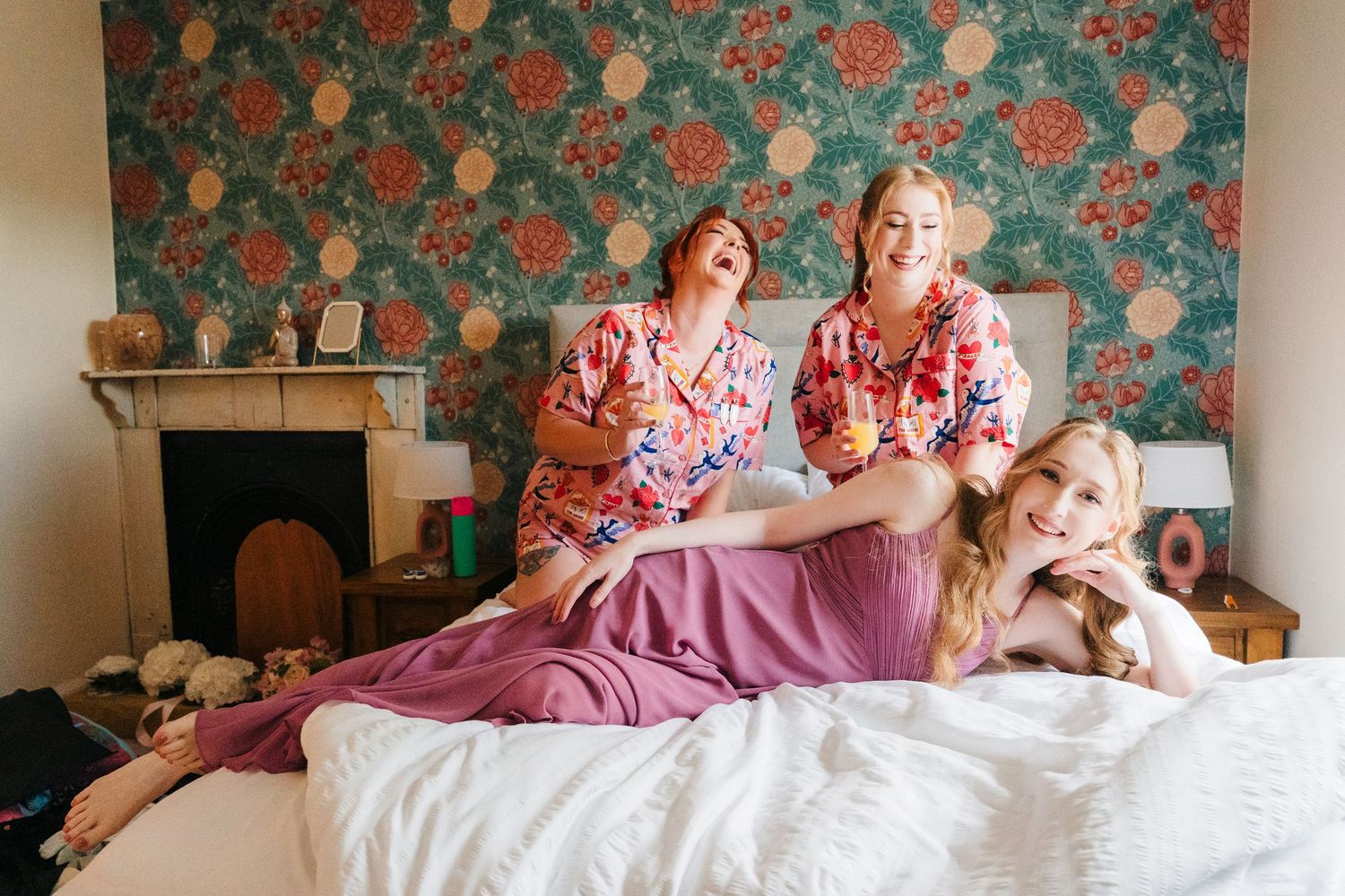 Three people in floral robes lounge together on a bed against vintage floral wallpaper with a fireplace in the background. . Village Barn Ty