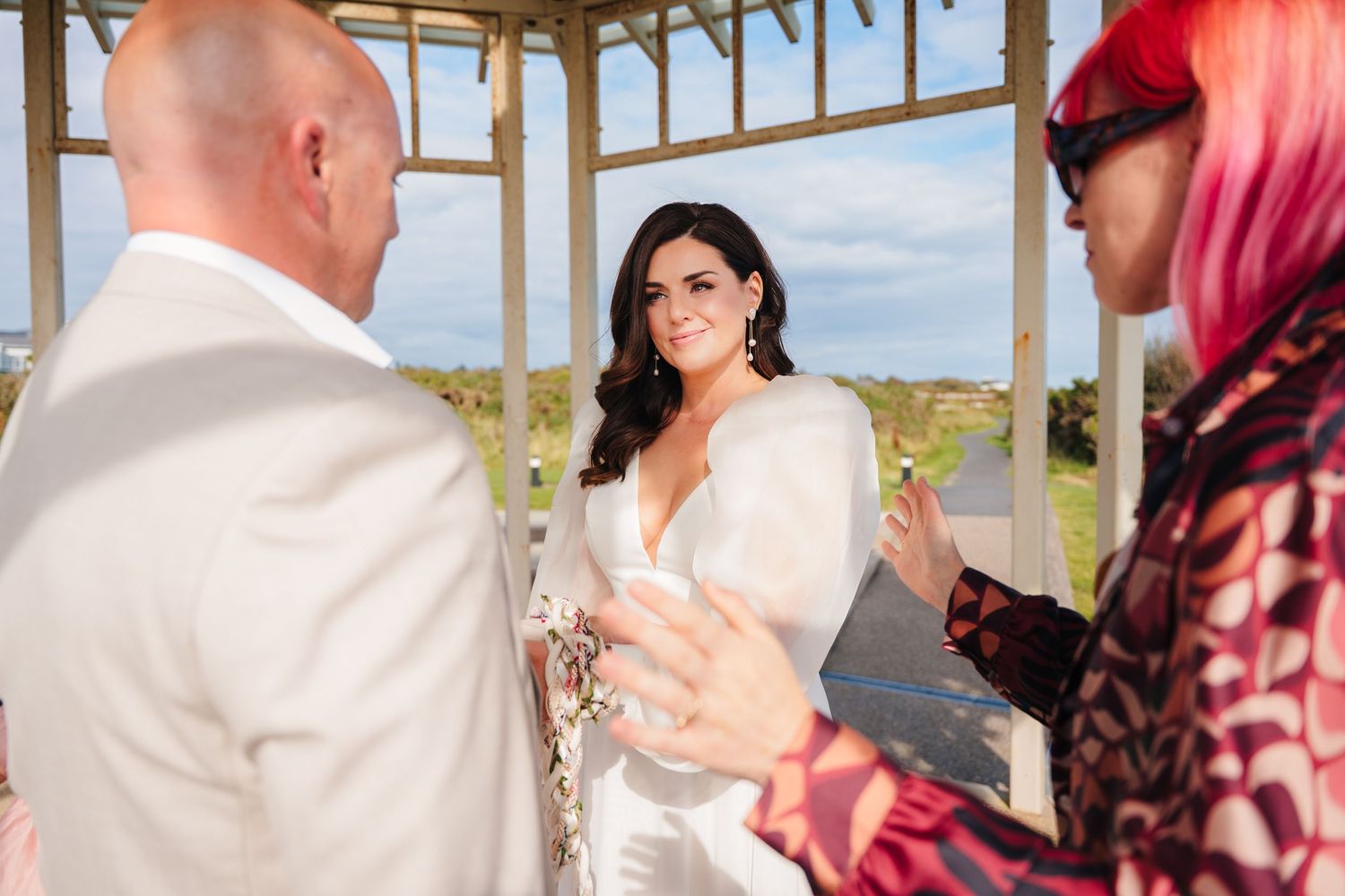 A couple exchanges vows during a beachside gazebo wedding ceremony with an officiant in vibrant attire.