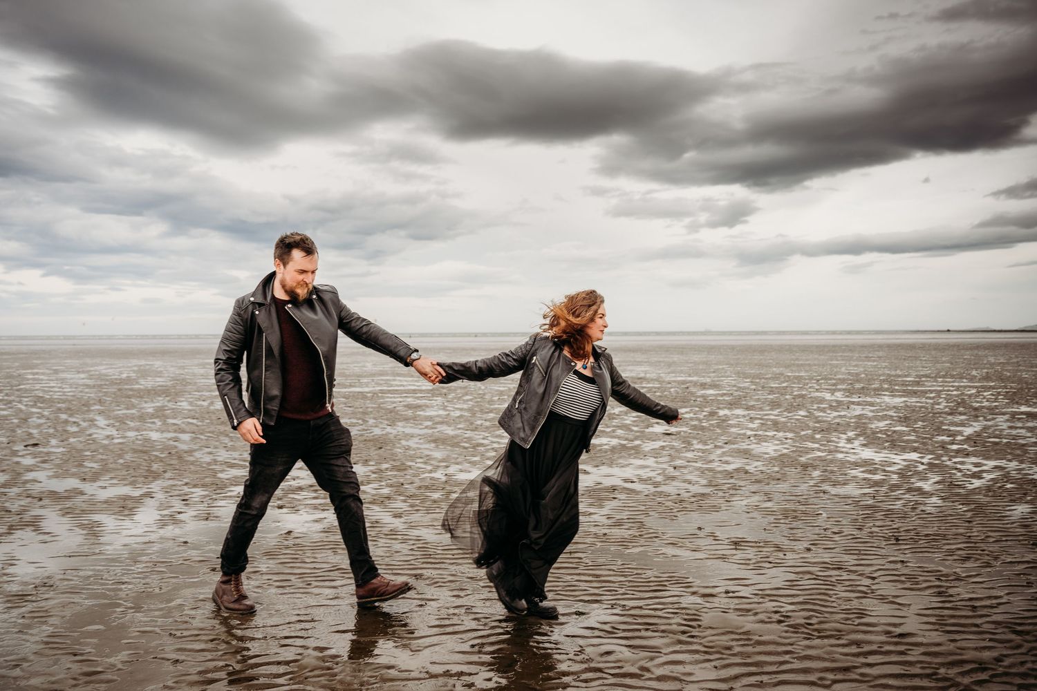 A couple dances and splashes playfully in the shallow ocean water under dramatic cloudy skies at sunset. engagement PHOTOGRAPHER Dublin