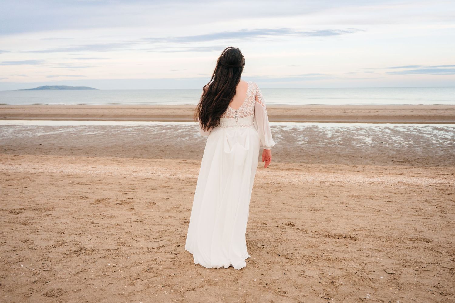 A woman in a flowing white dress stands alone on a beach at sunset looking towards the horizon. Portmarnock Hotel Wedding PHOTOGRAPHER Dubli
