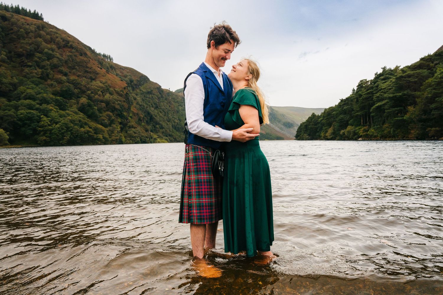 A couple embraces romantically by a misty lake surrounded by mountains while wearing traditional Scottish attire.Family photogrAPHER DUBLIN