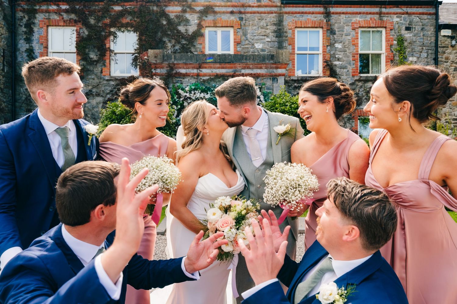 Wedding photographer DUBLIN couple kissing surrounded by bridesmaids in pink dresses and groomsmen in navy suits holding bouquets outdoors.