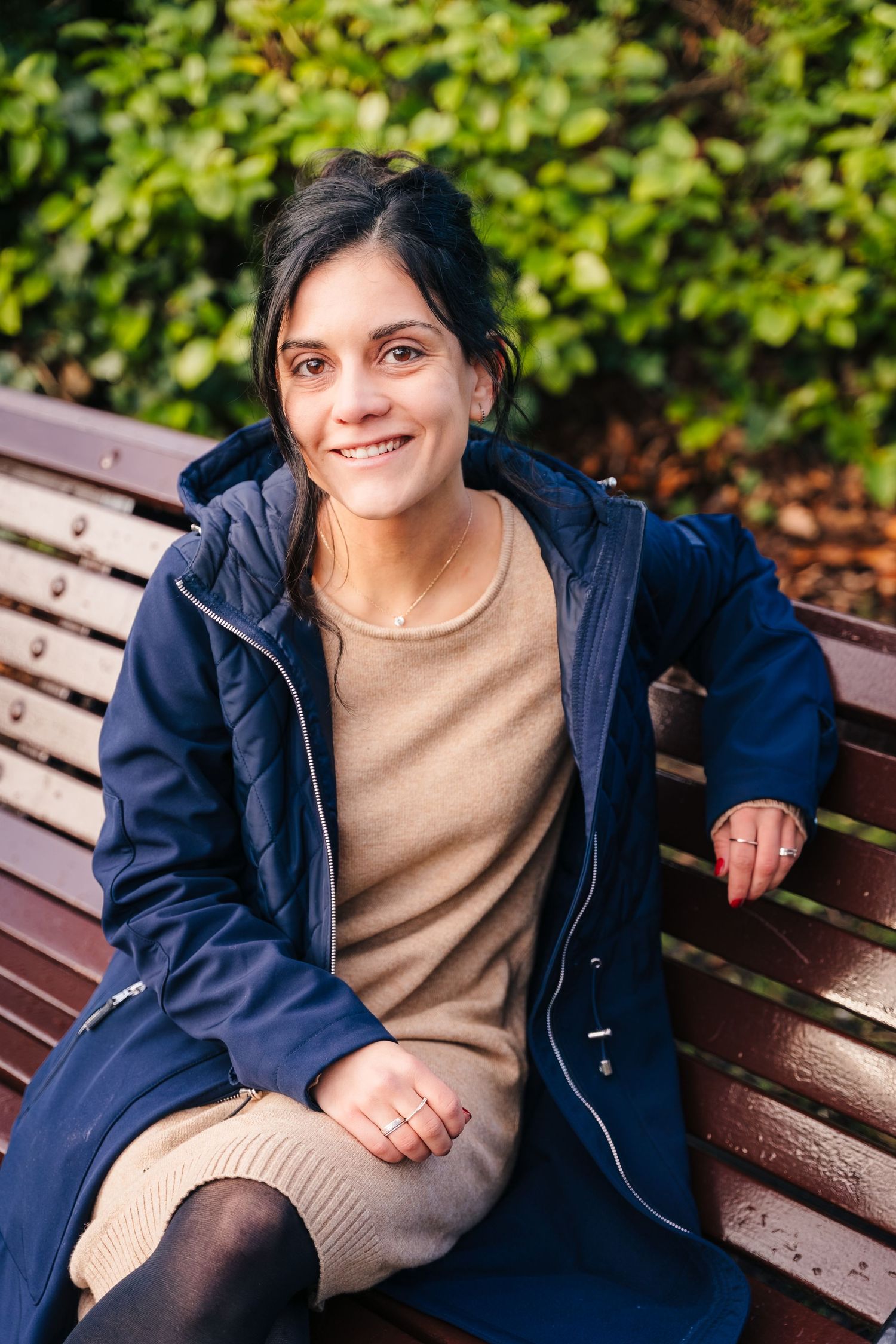 Person in navy jacket and beige sweater sitting on wooden park bench with greenery in background. professional headshots dublin