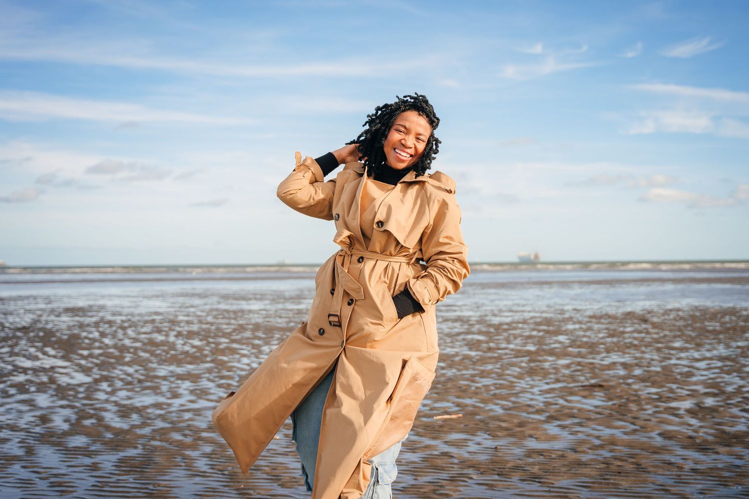 A joyful fashion shoot on a beach featuring a tan trench coat against a bright blue sky and sandy shore. Dublin professional headshots