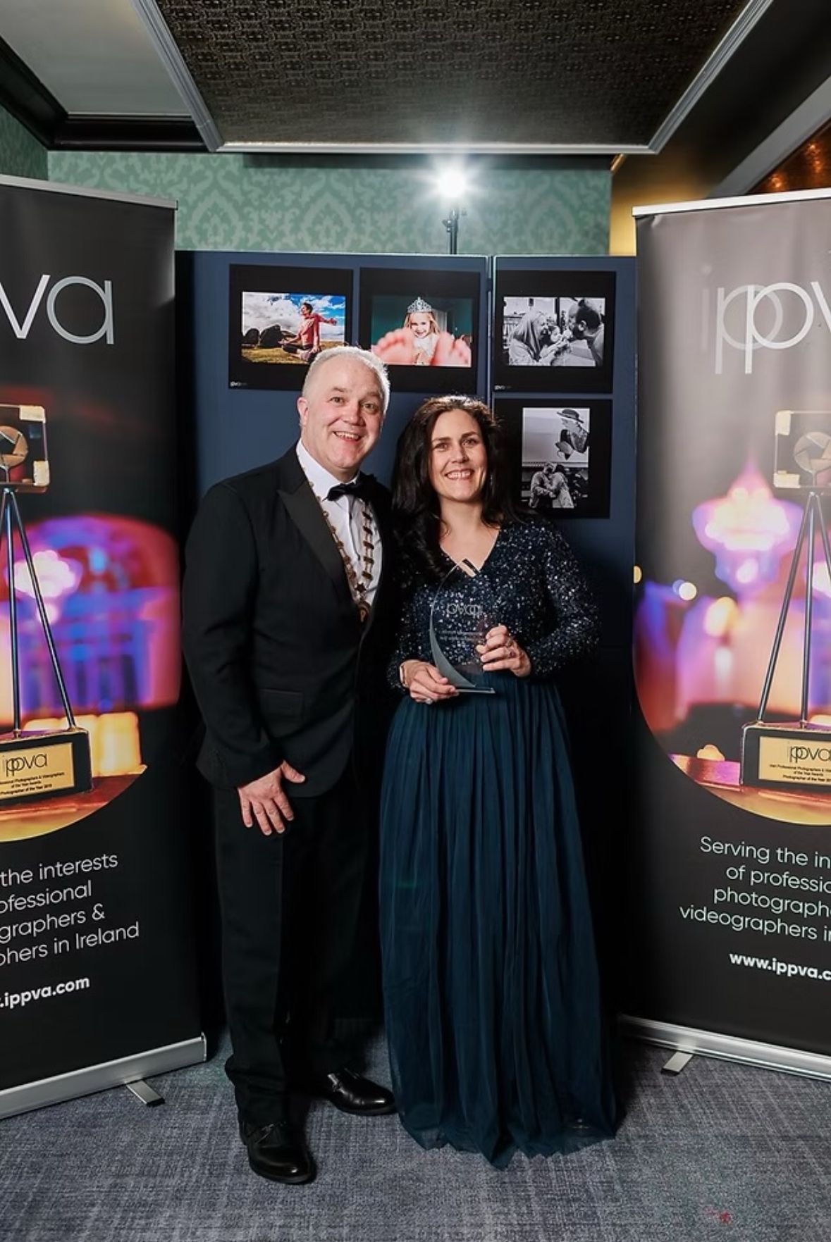 A couple poses with award banners at a photography exhibition showing mounted prints and displays.