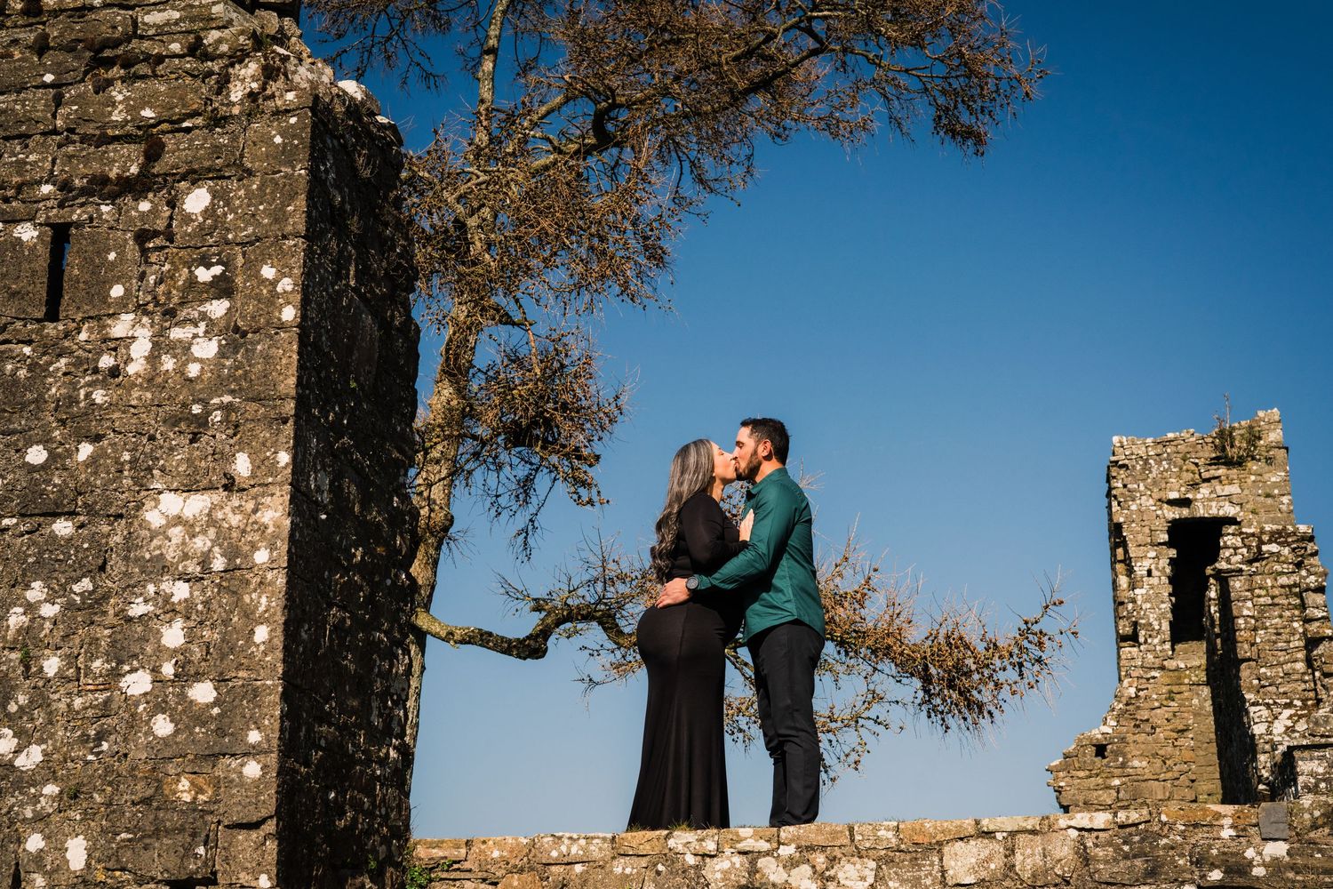 A couple takes romantic engagement photos at golden hour against ancient stone ruins and blue sky backdrop. PHOTOGRAPHER Dublin ireland