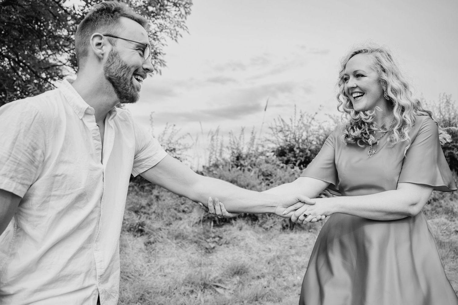 Black and white photo of a joyful couple laughing together while holding hands outside in a natural setting. PHOTOGRAPHER Dublin ireland