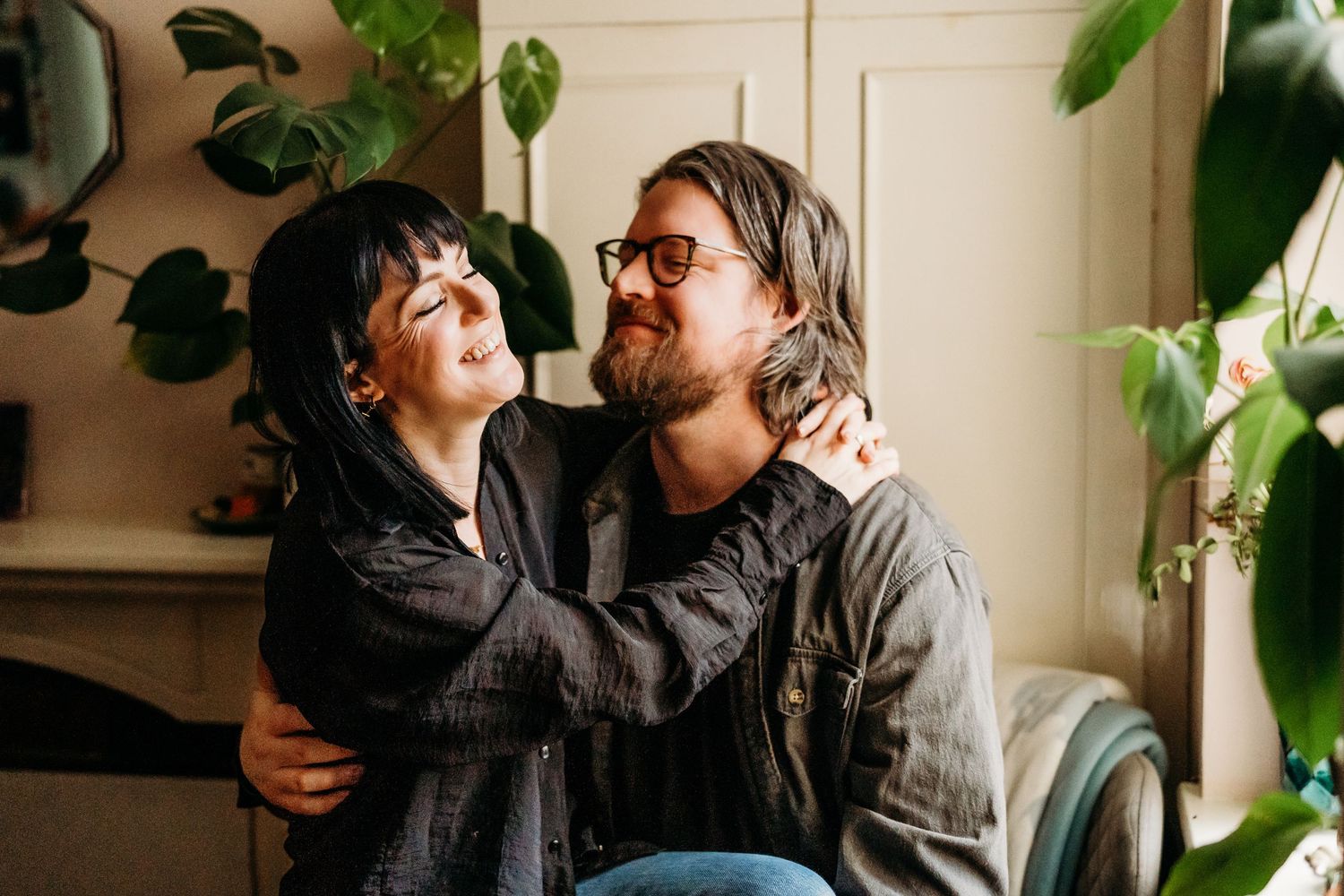 A couple shares a tender moment and laughter while sitting together in a sunlit room with houseplants in the background. PHOTOGRAPHER Dublin