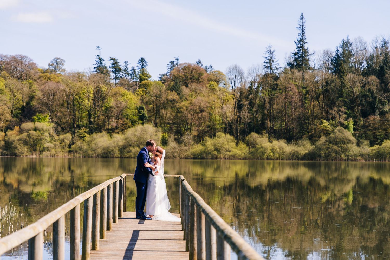 Newlywed couple embracing on a wooden boardwalk overlooking a serene lake surrounded by autumn trees. Wedding PHOTOGRAPHER Dublin