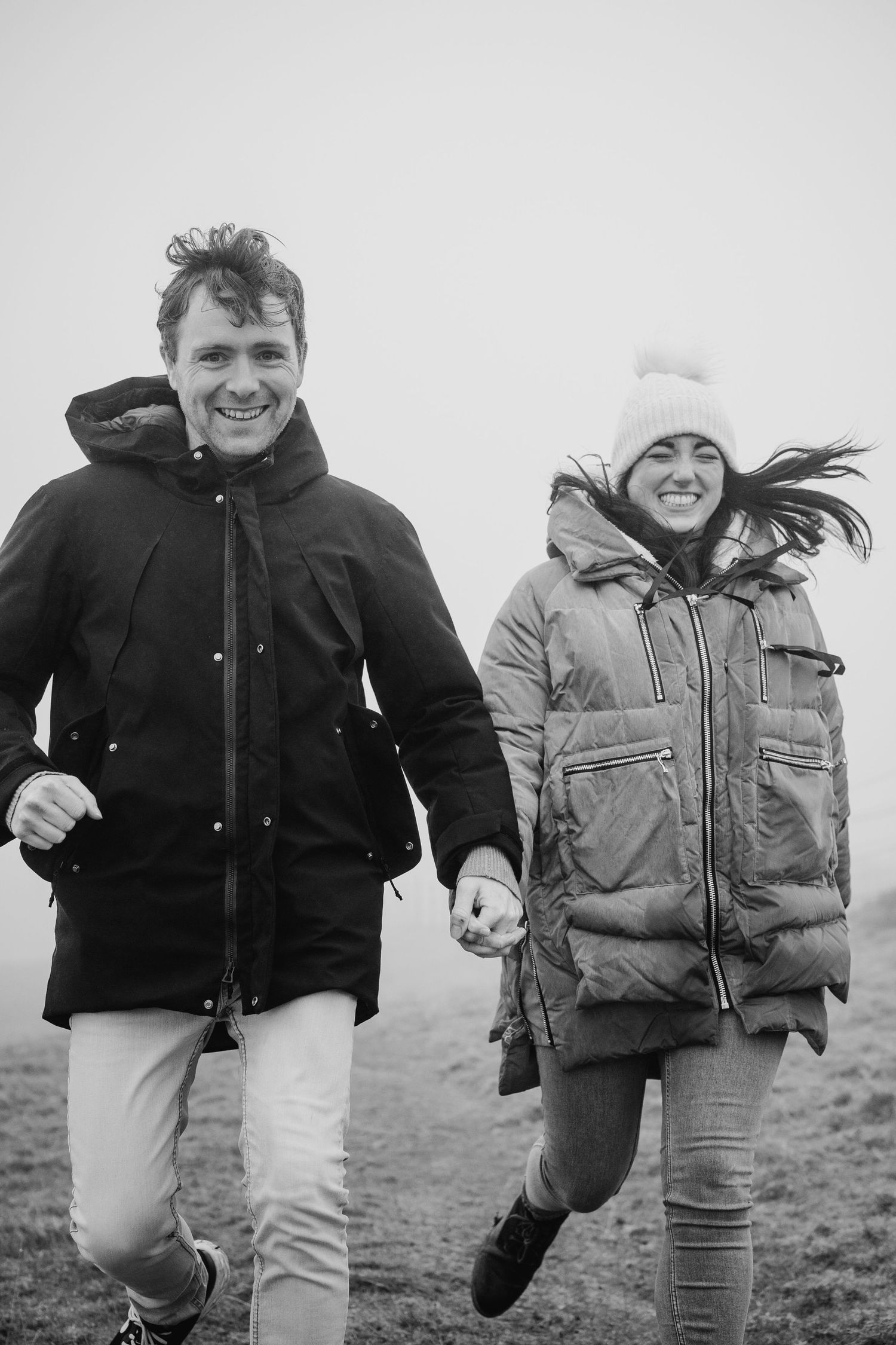 Black and white photo of two people in winter coats running and laughing together on a misty day outdoors.