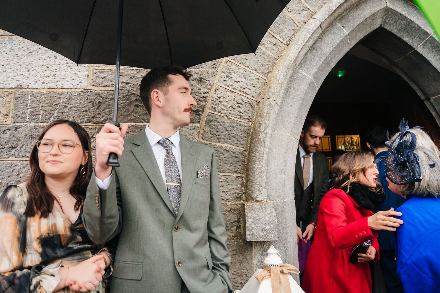 Wedding guests sharing joyful moments under umbrellas outside a stone church building.