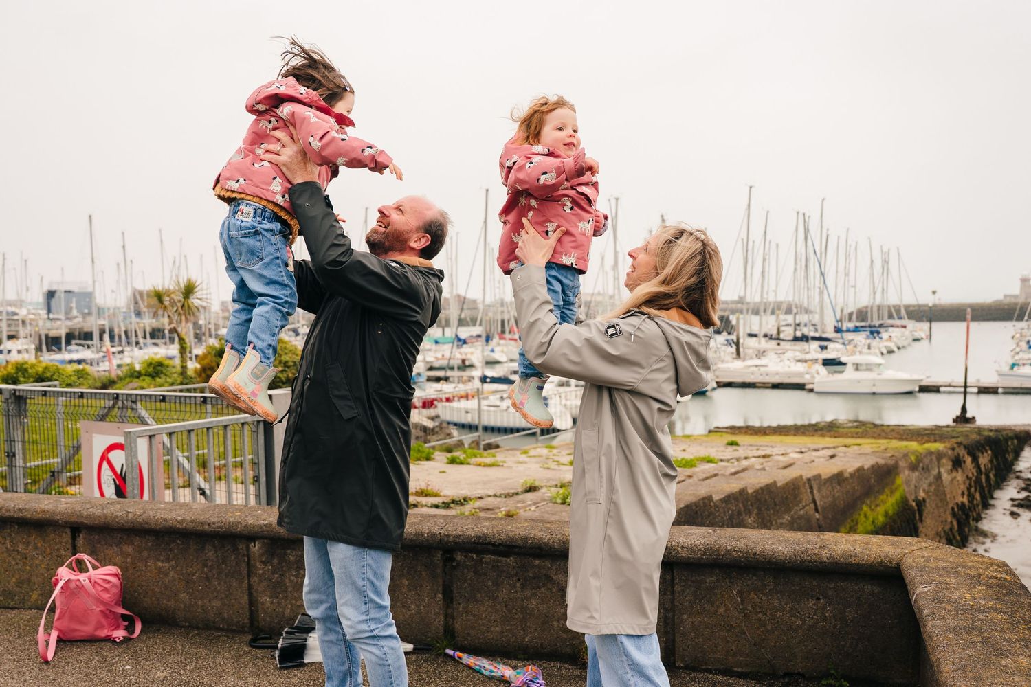 Adults playfully lift small children wearing pink jackets by a marina with boats in the background. PHOTOGRAPHER Dublin