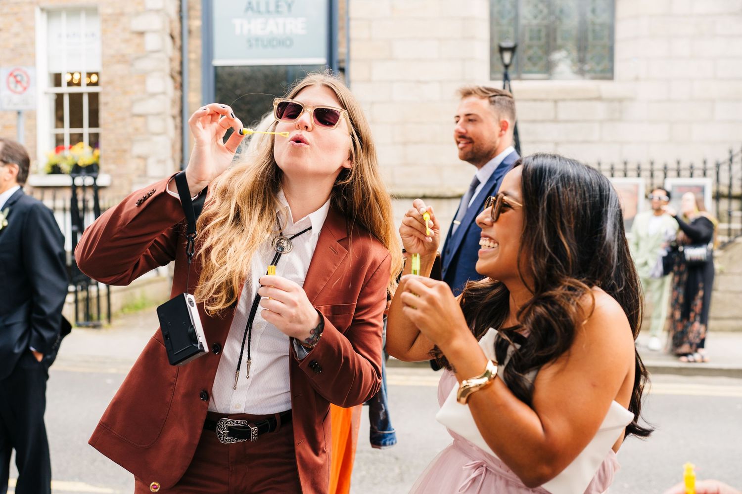 Wedding guests socialize and take photos on a sunny city street. PHOTOGRAPHER Dublin Ireland Wedding