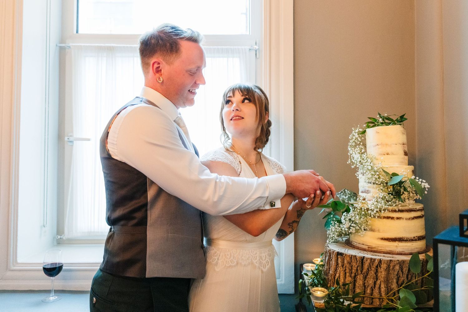 Couple cuts their rustic three-tiered wedding cake decorated with greenery near a window. PHOTOGRAPHER Dublin Ireland Wedding