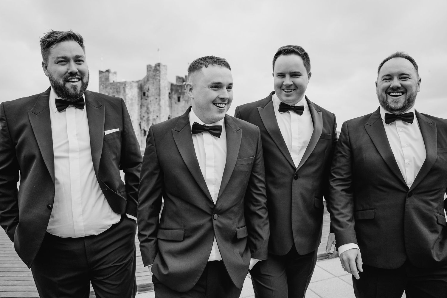 A group of well-dressed groomsmen in black tuxedos and bowties laugh together while standing in a line outdoors. Wedding Photography Trim Ca