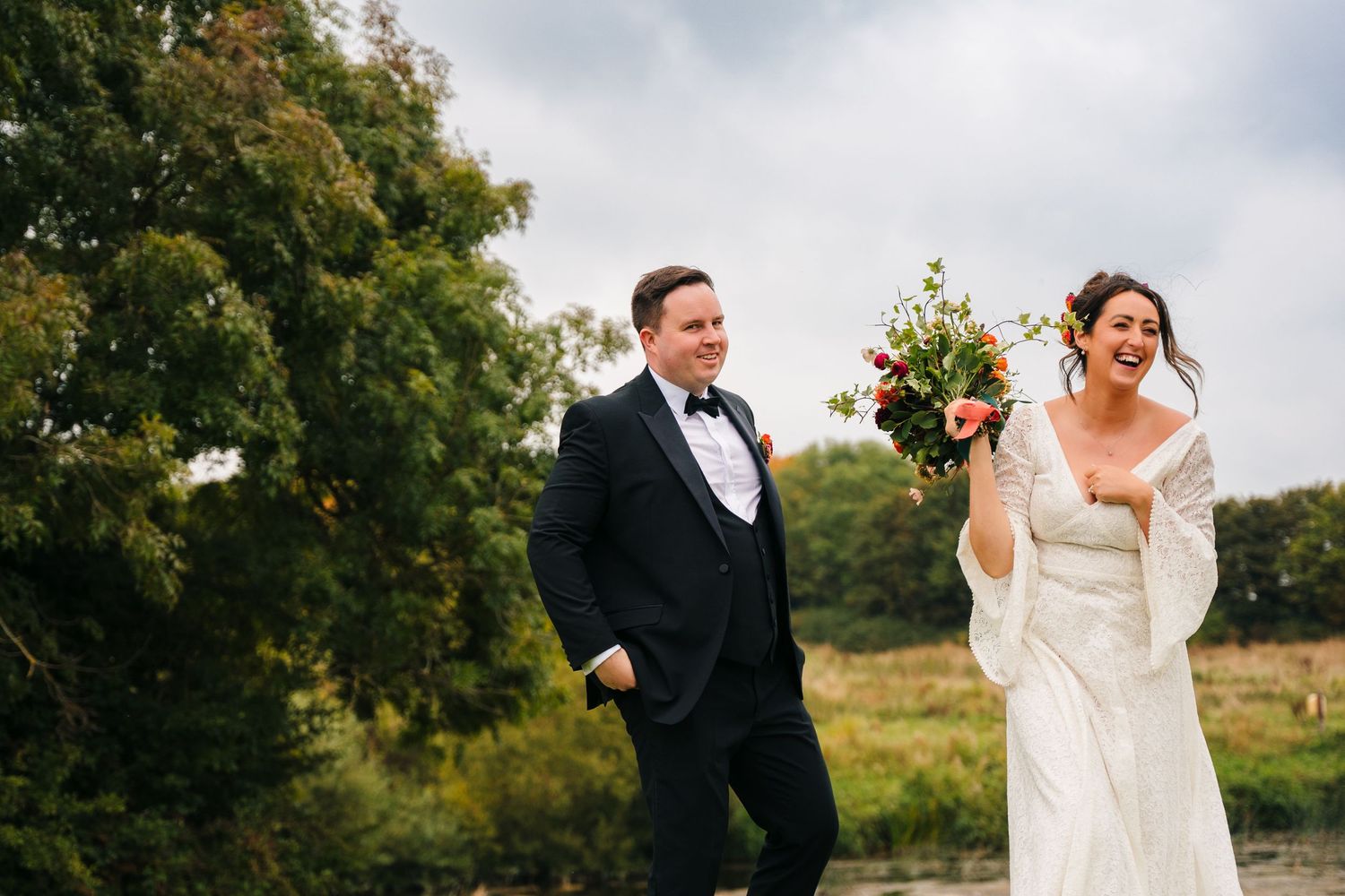 Newlyweds playfully running and laughing together in a green field under cloudy skies.Wedding Photography Trim Castle Ireland