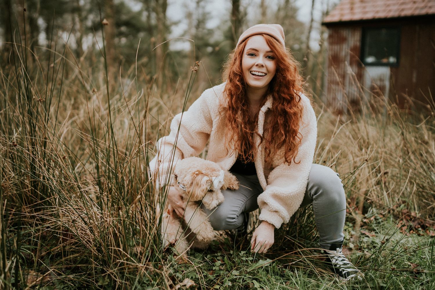 A person with long red hair crouches in tall grass wearing a cozy fur jacket while smiling outdoors near a rustic cabin. PHOTOGRAPHER Dublin