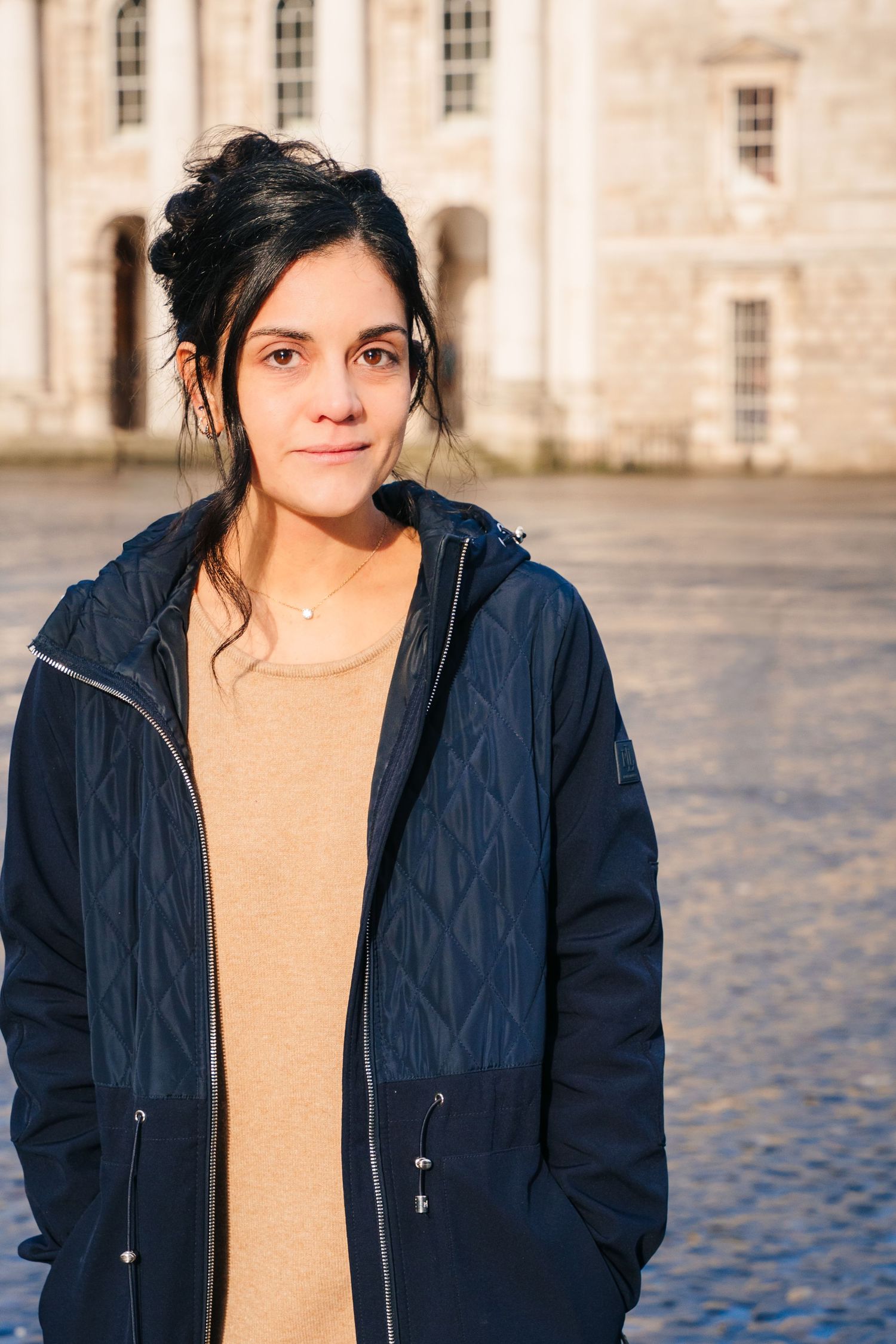 A person wearing a navy blue quilted jacket and tan top stands in front of a historic stone building on a sunny day. professional headshots