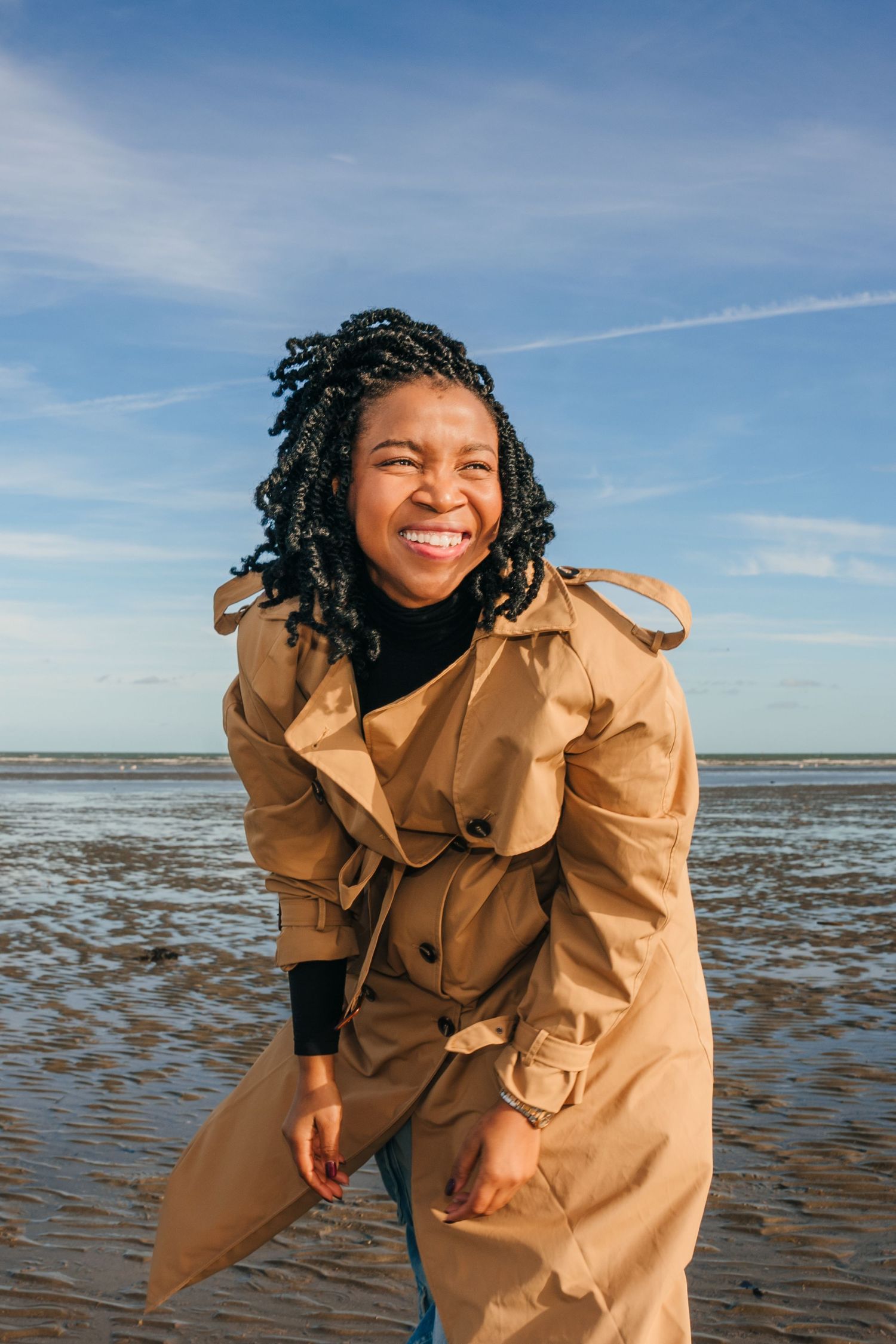 Person wearing tan trench coat laughing on beach with bright blue sky background. Dublin professional headshots photographer