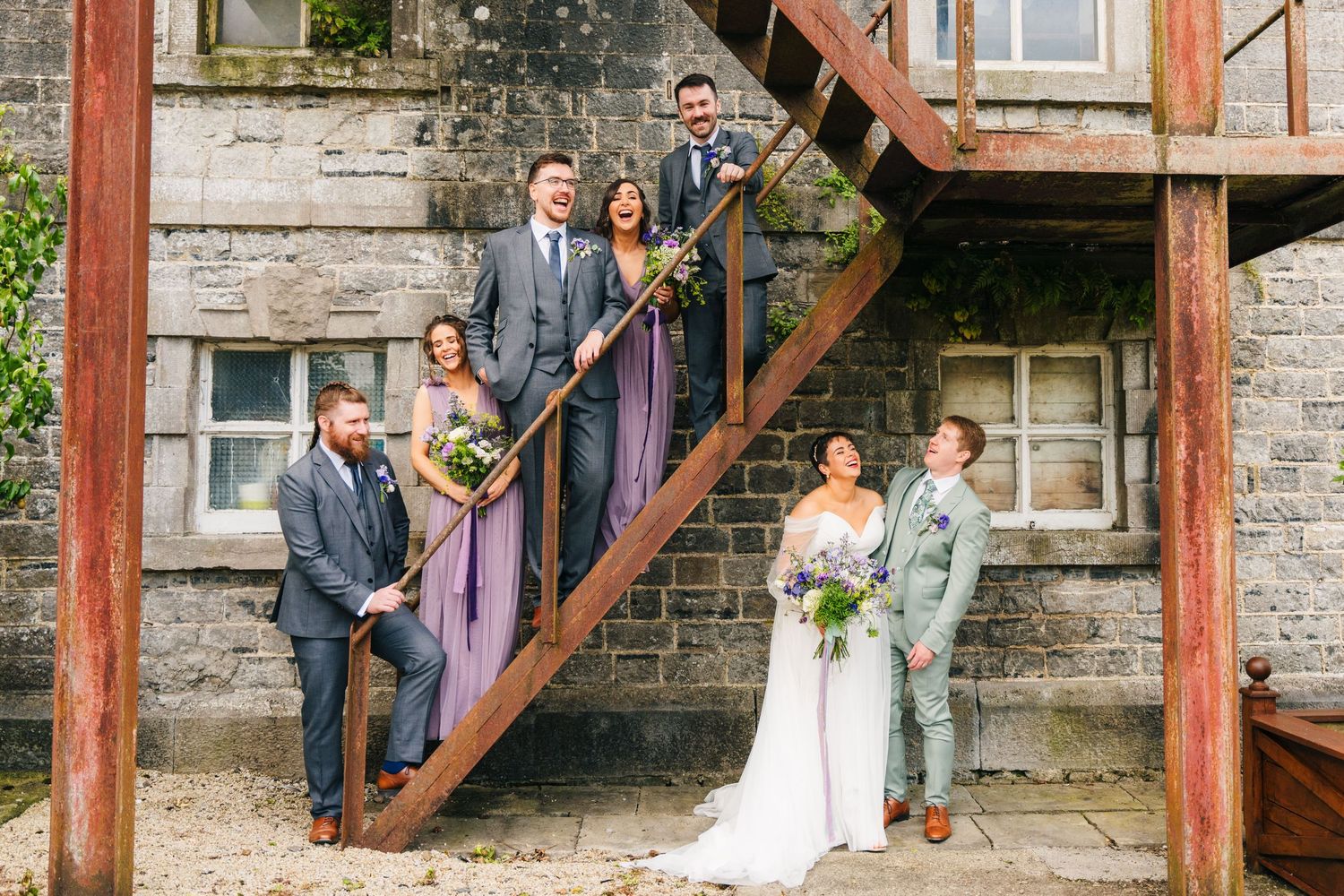 Wedding party poses on rustic wooden staircase against stone wall backdrop during outdoor celebration.