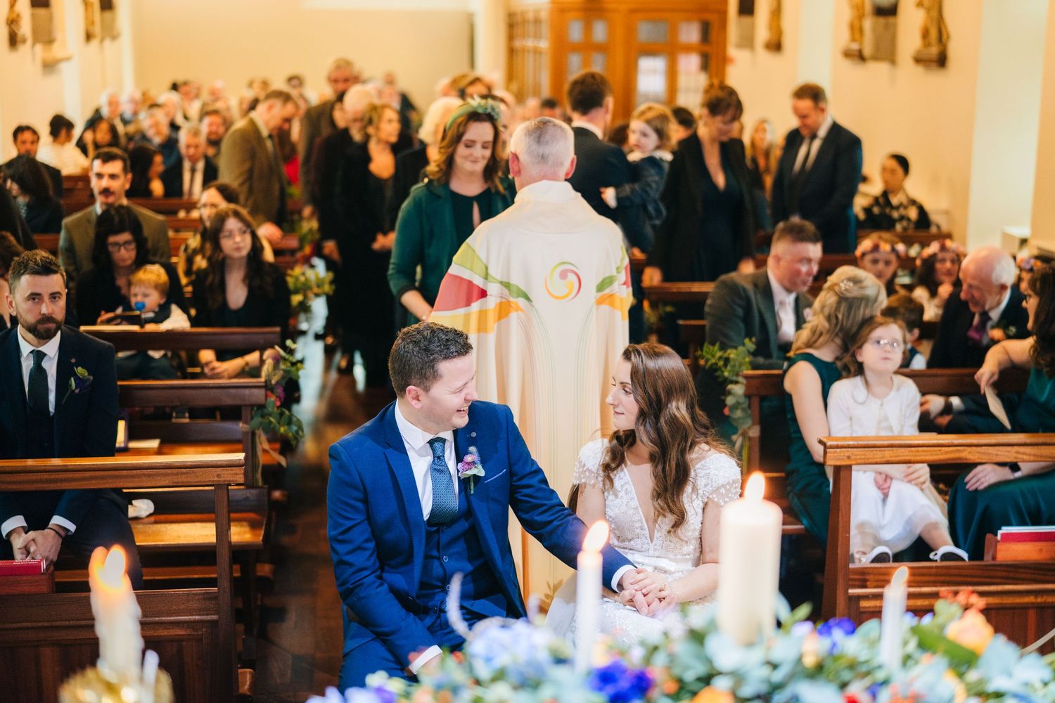 Guests watching a wedding ceremony from wooden pews in a sunlit church.
